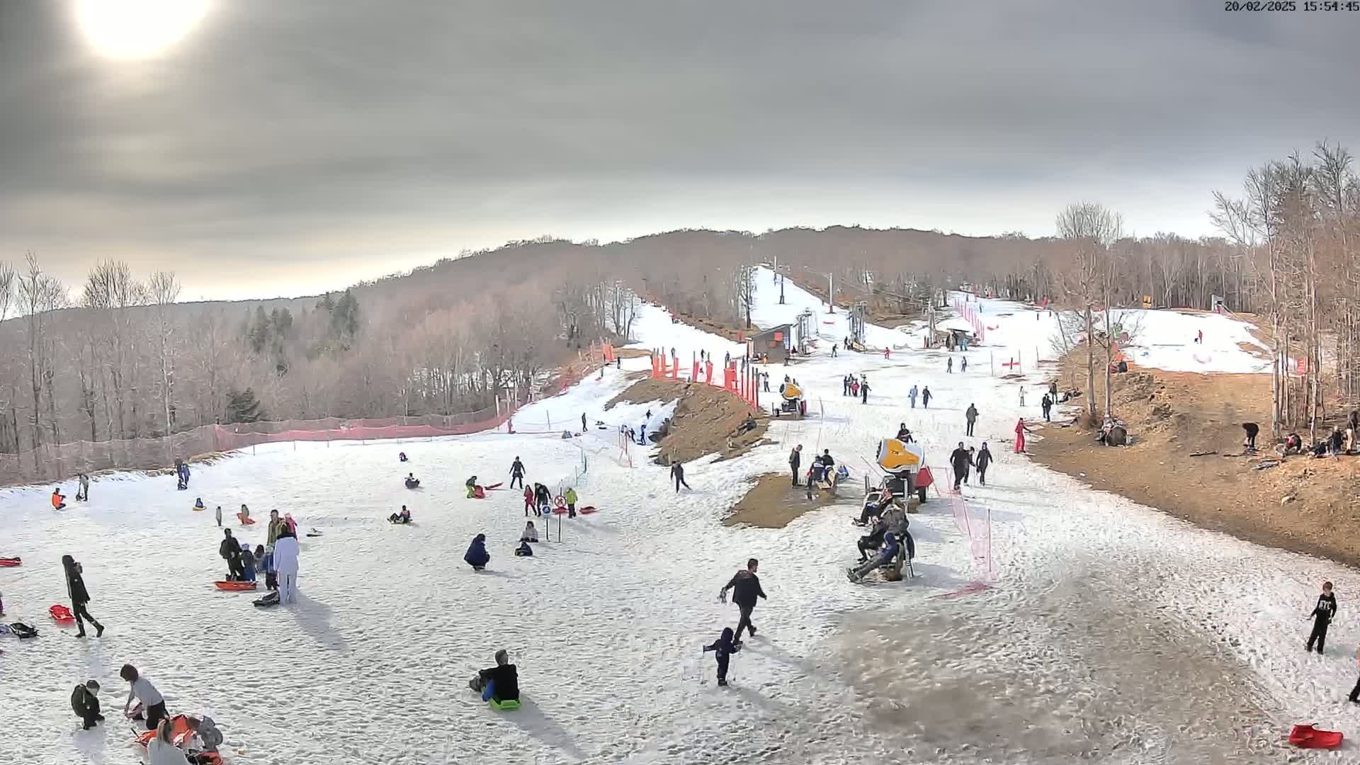 A snowy hill with a ski lift and many people sledding and walking under a partly sunny sky.