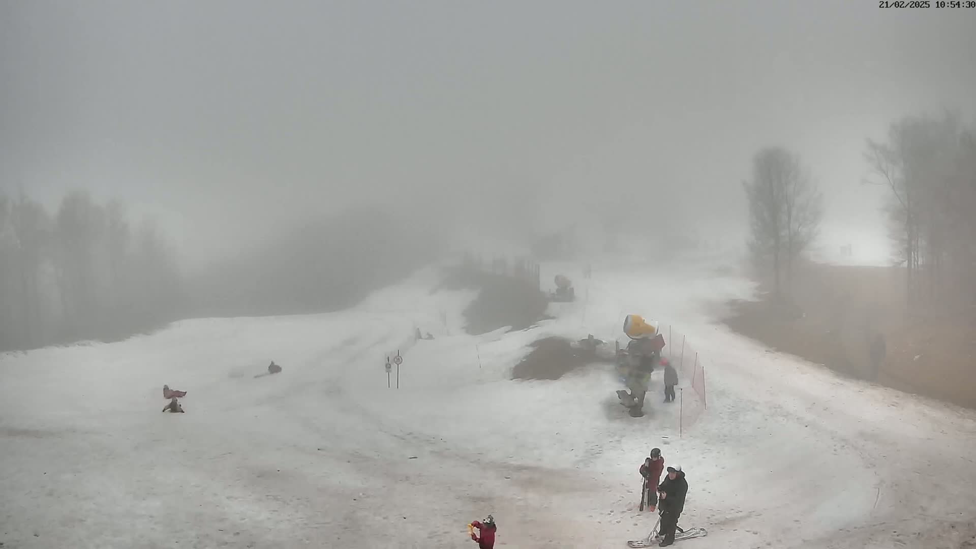 A snow-covered ski slope is shrouded in thick fog, with a few people skiing and snowboarding on the hill.