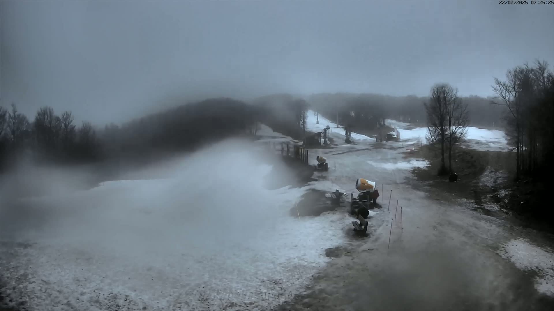 Snow cannons are making snow on a ski slope under an overcast sky.
