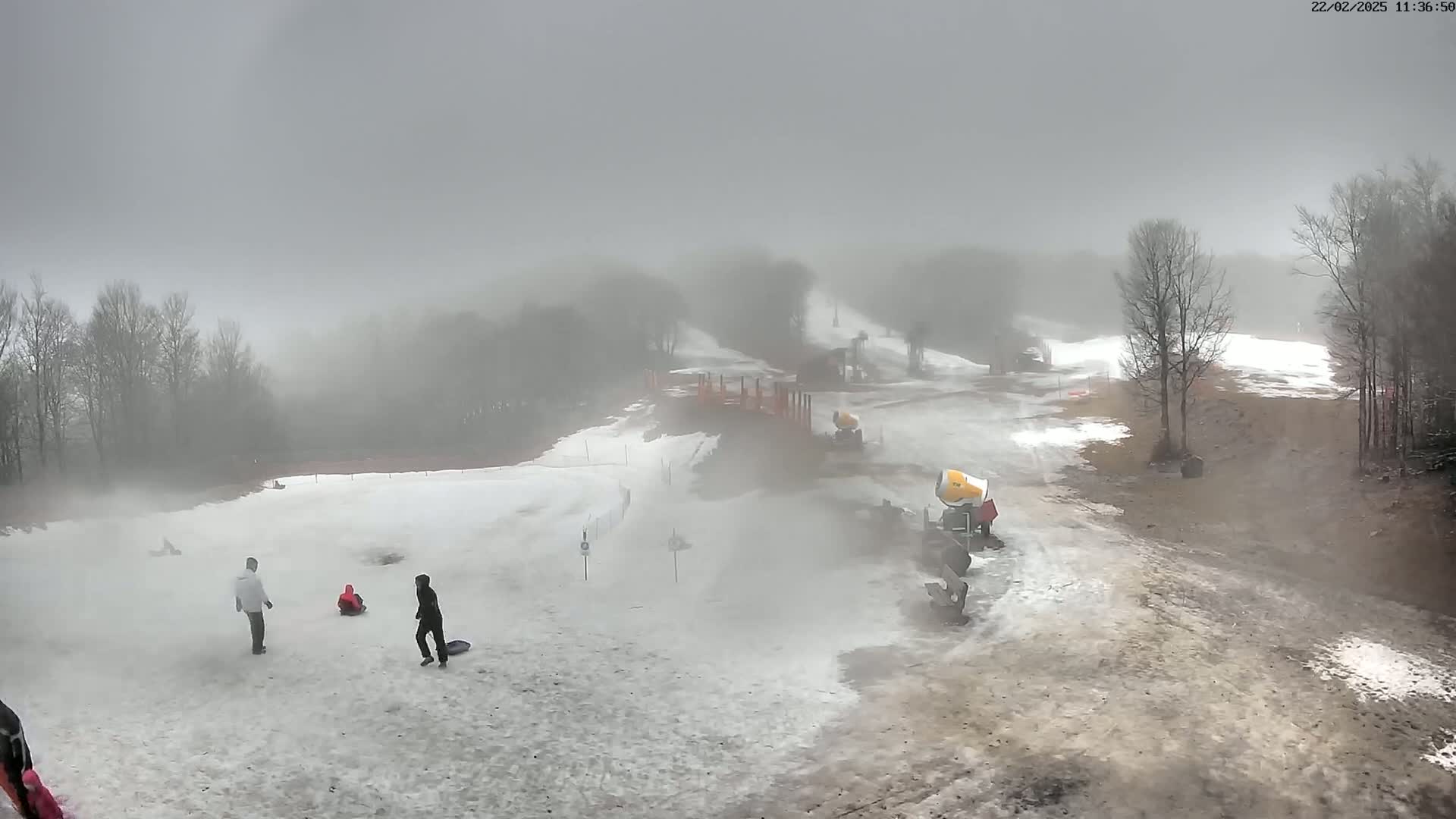 A snowy, foggy scene shows a few people sledding on a hill, with snowmaking machines visible in the background.