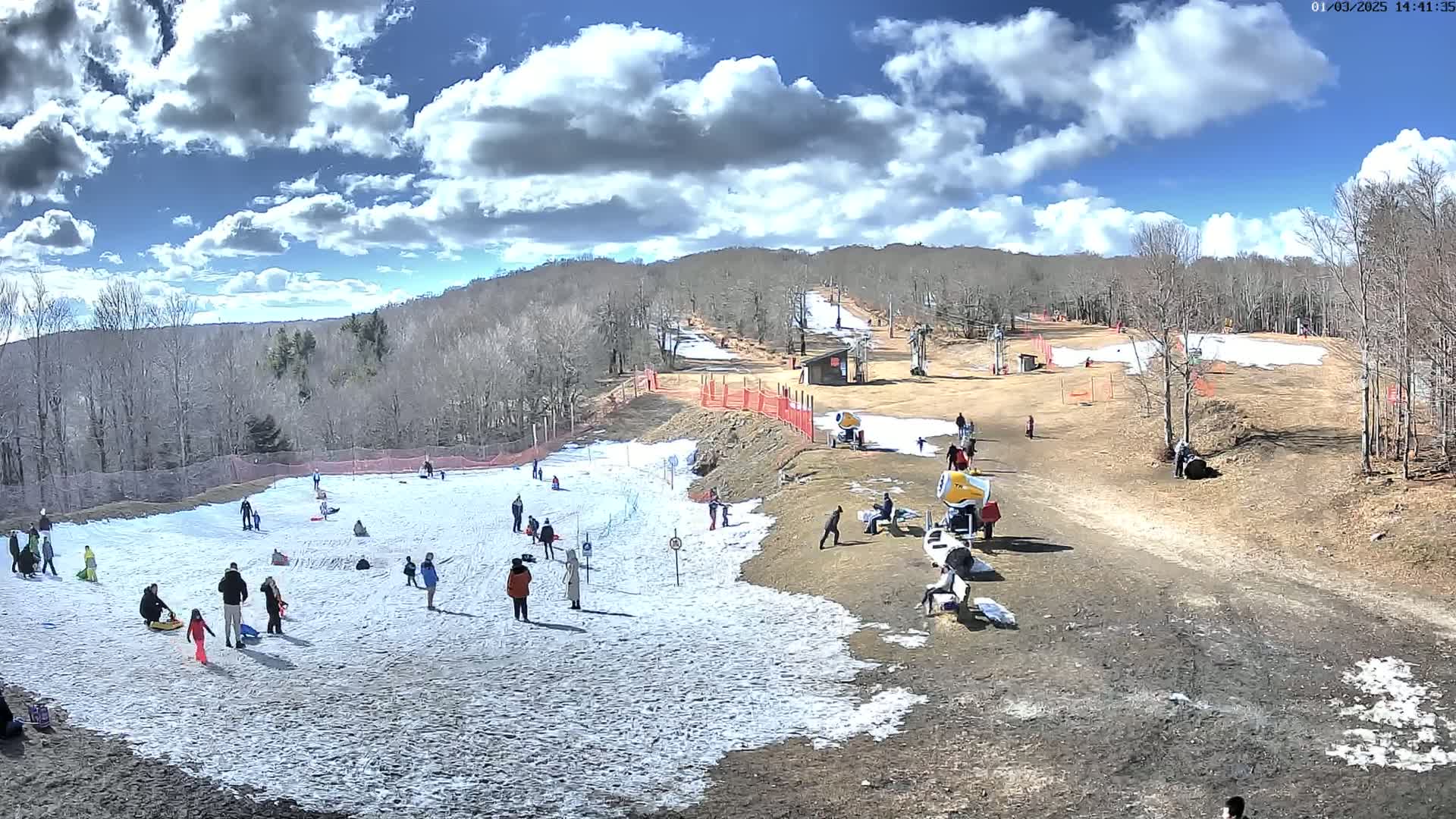 A partly sunny day shows a snow-covered hill with people sledding and snowmaking equipment, surrounded by bare trees and brown ground.