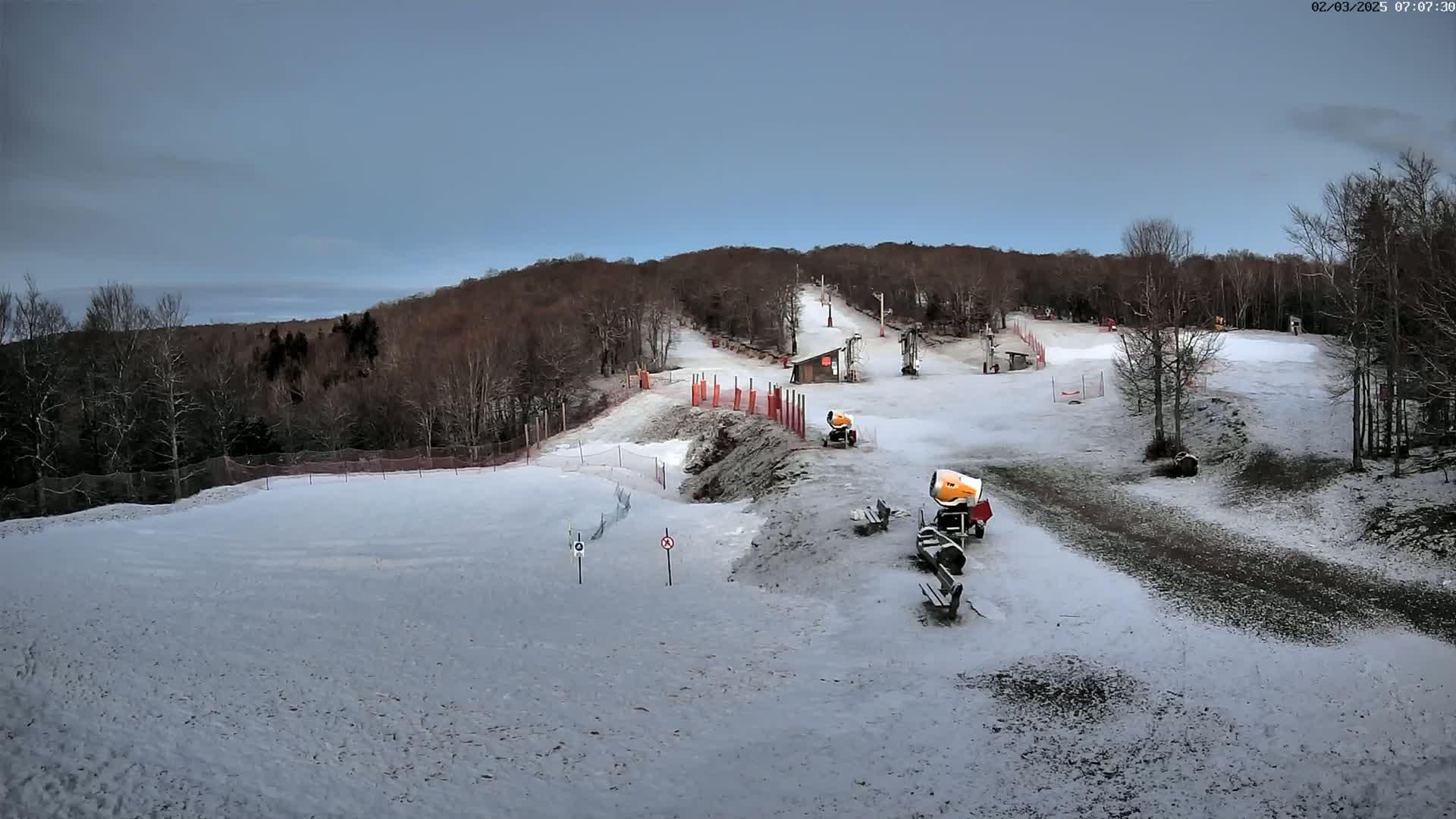 A snow-covered ski slope with snow-making machines, a ski lift, and bare deciduous trees under a cloudy sky.