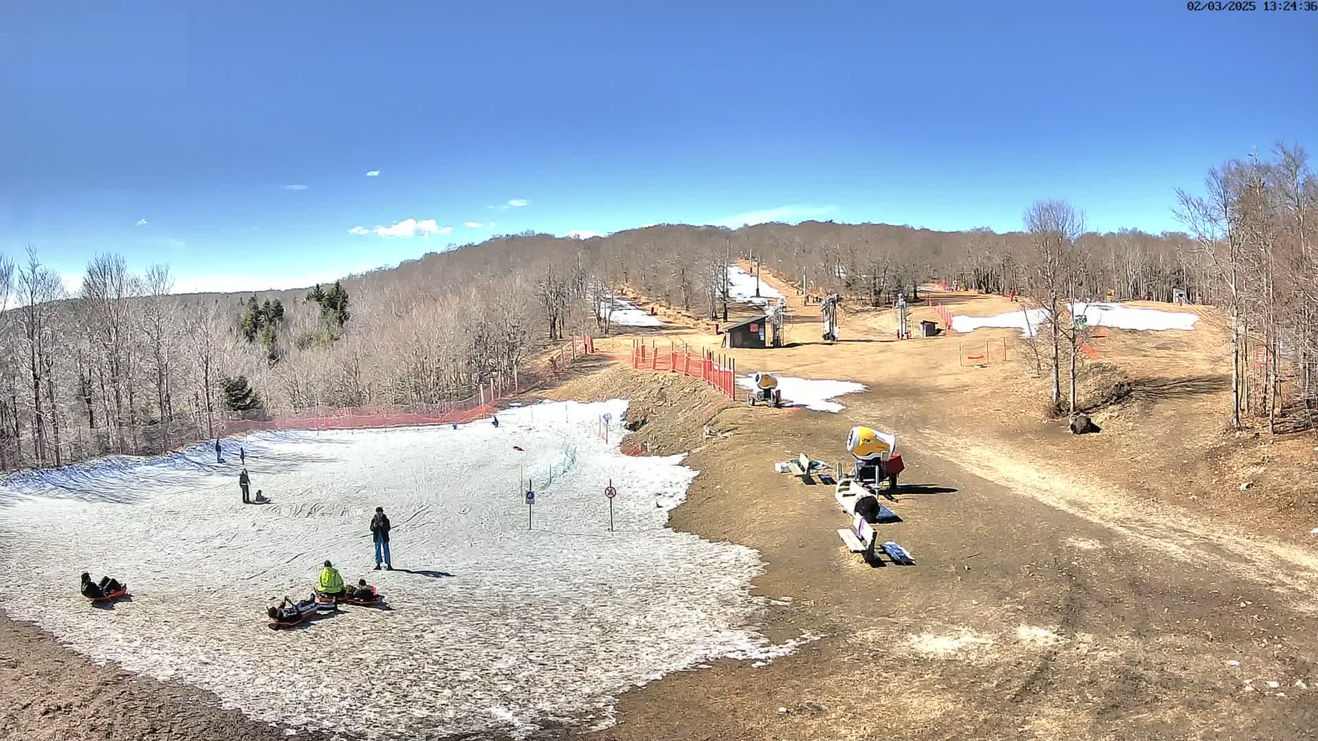 A mostly sunny day at a snow tubing hill shows several people tubing down a small, partially snow-covered slope, with snowmaking machines and a ski lift visible in the background.