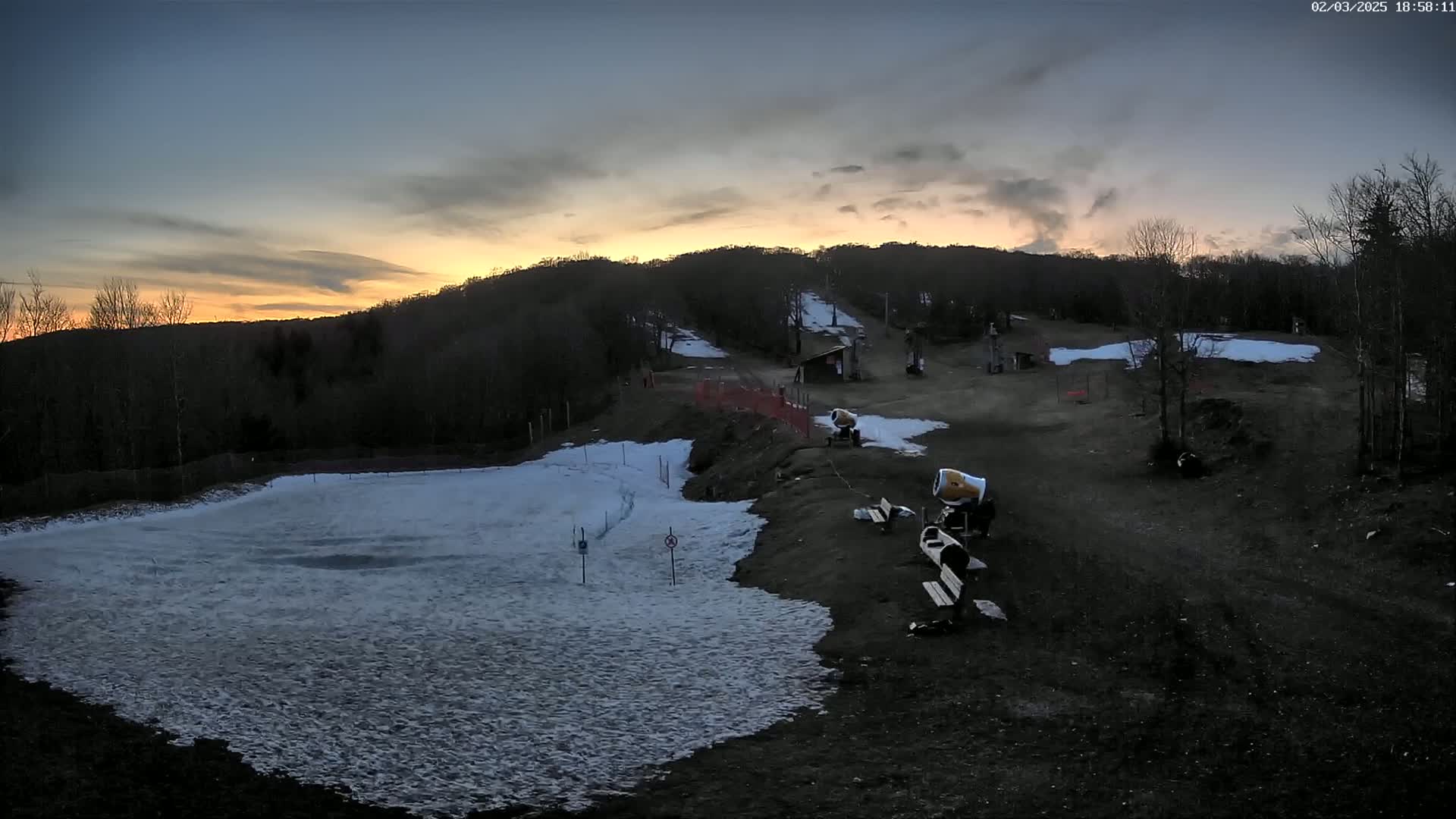 A partially snow-covered ski slope at sunset, with snowmaking equipment present.