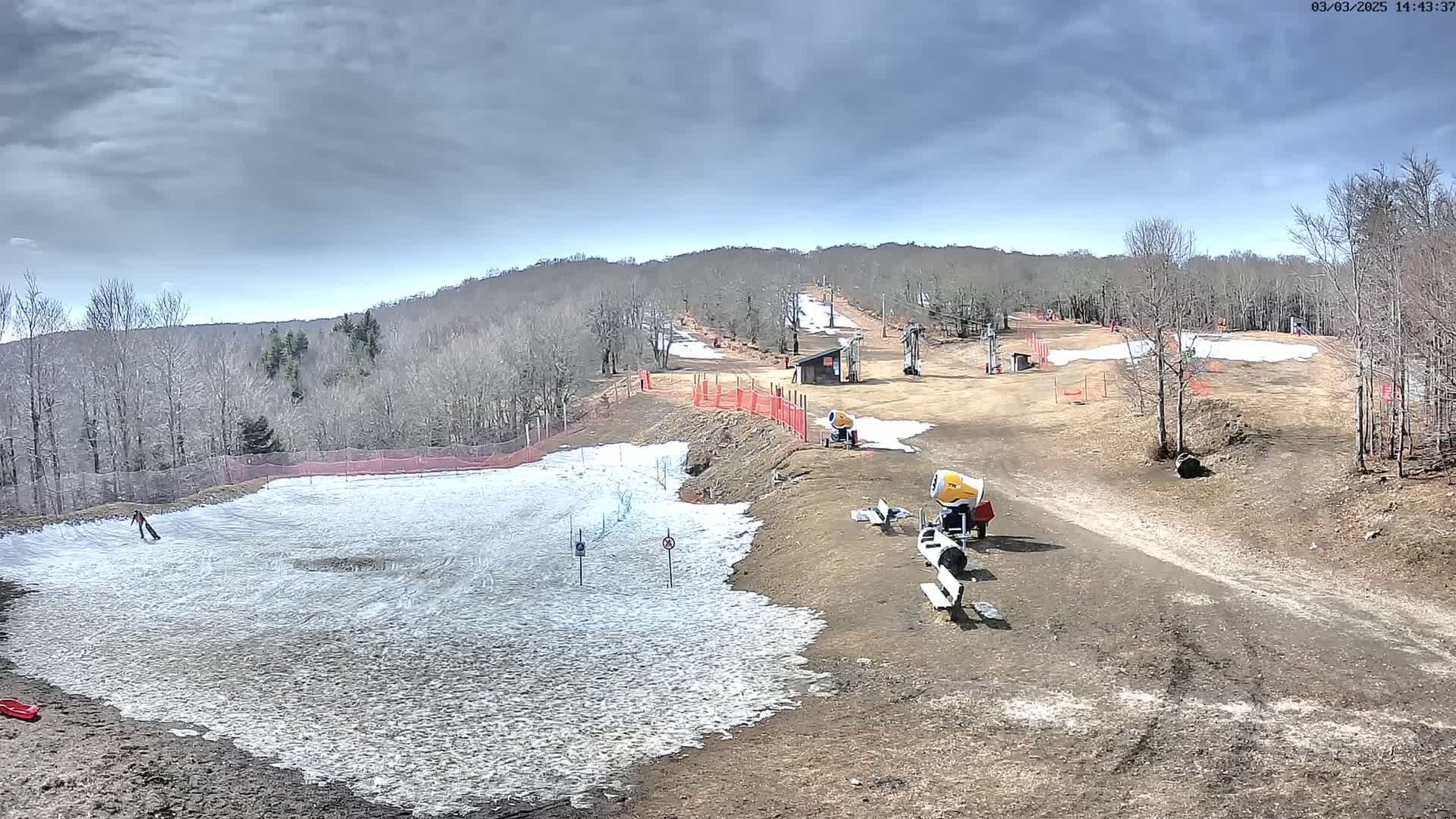 A partially snow-covered ski slope with snowmaking machines, under a mostly cloudy sky.