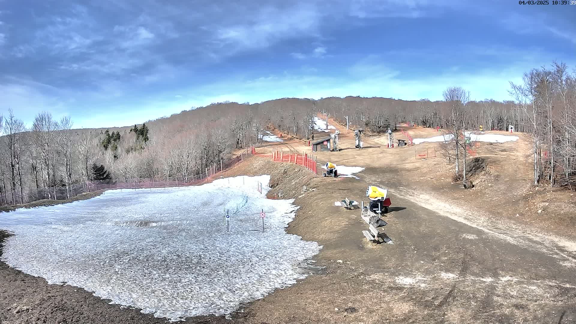 A mostly barren ski slope with patches of snow, snowmaking machines, and a chairlift under a partly cloudy sky.