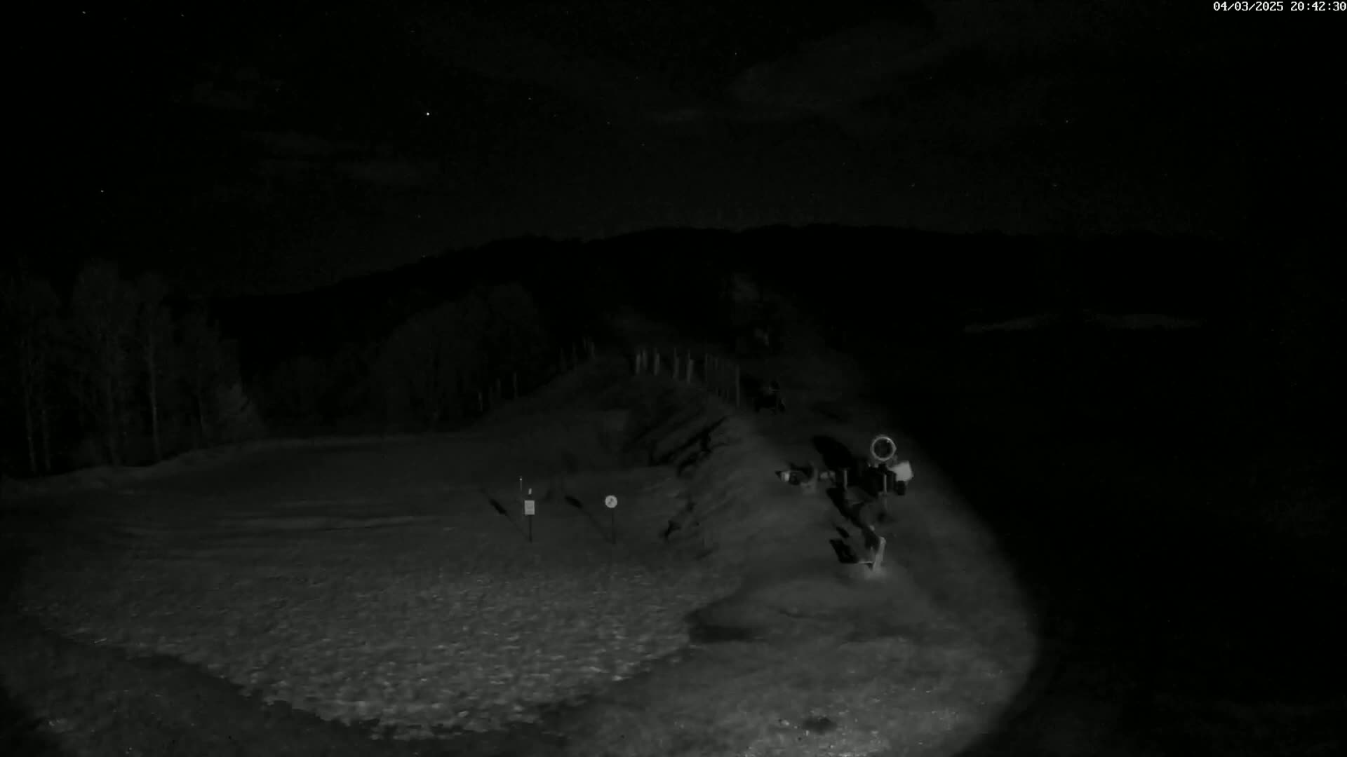 A nighttime, snow-covered area is illuminated by a light source, revealing a small structure and some trees in the background under a partly cloudy sky.