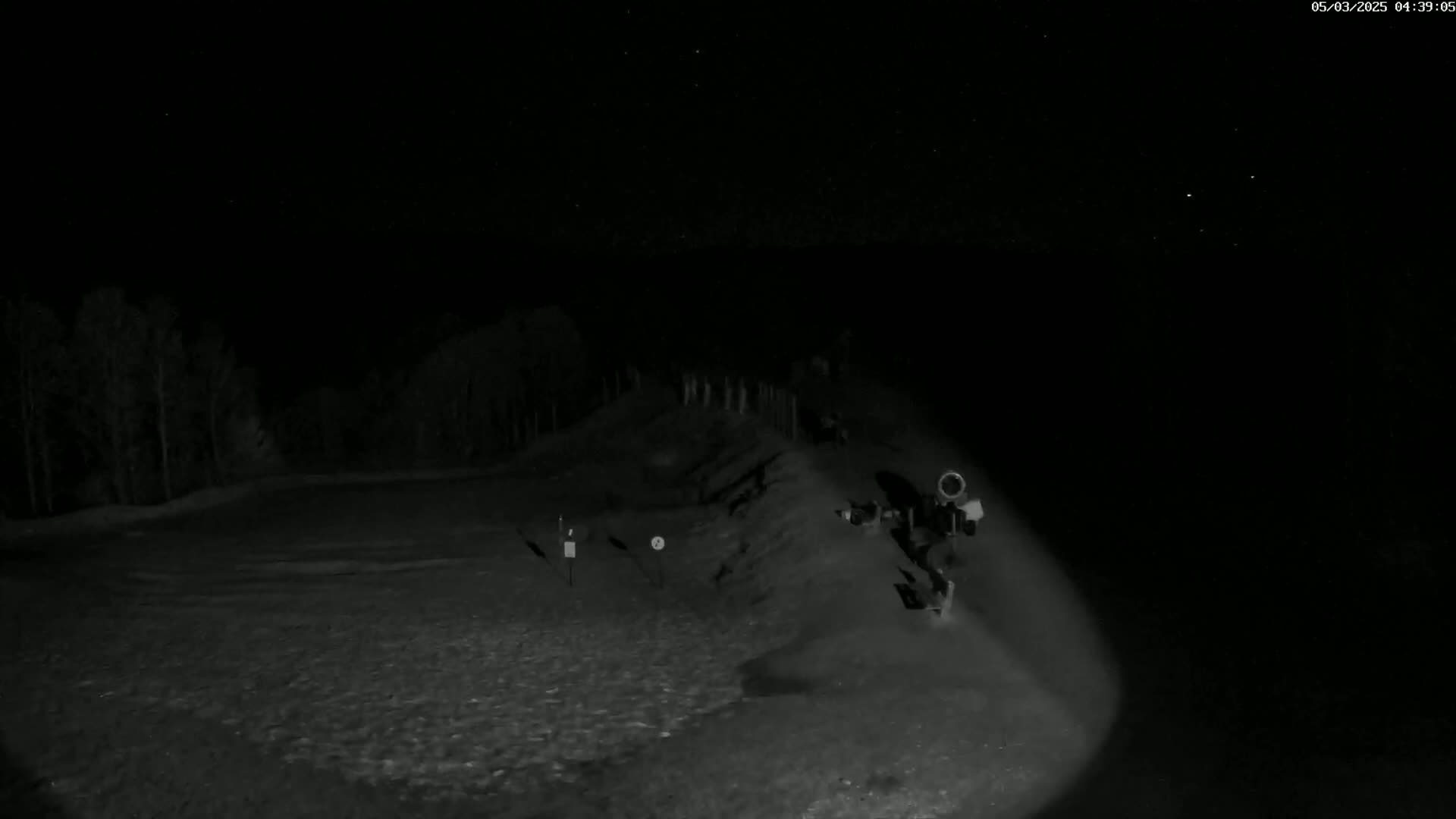 A nighttime, low-light image shows a dark, mostly barren landscape with a few trees in the background, a small mound in the middle ground, and what appears to be a camera or monitoring equipment in the foreground.