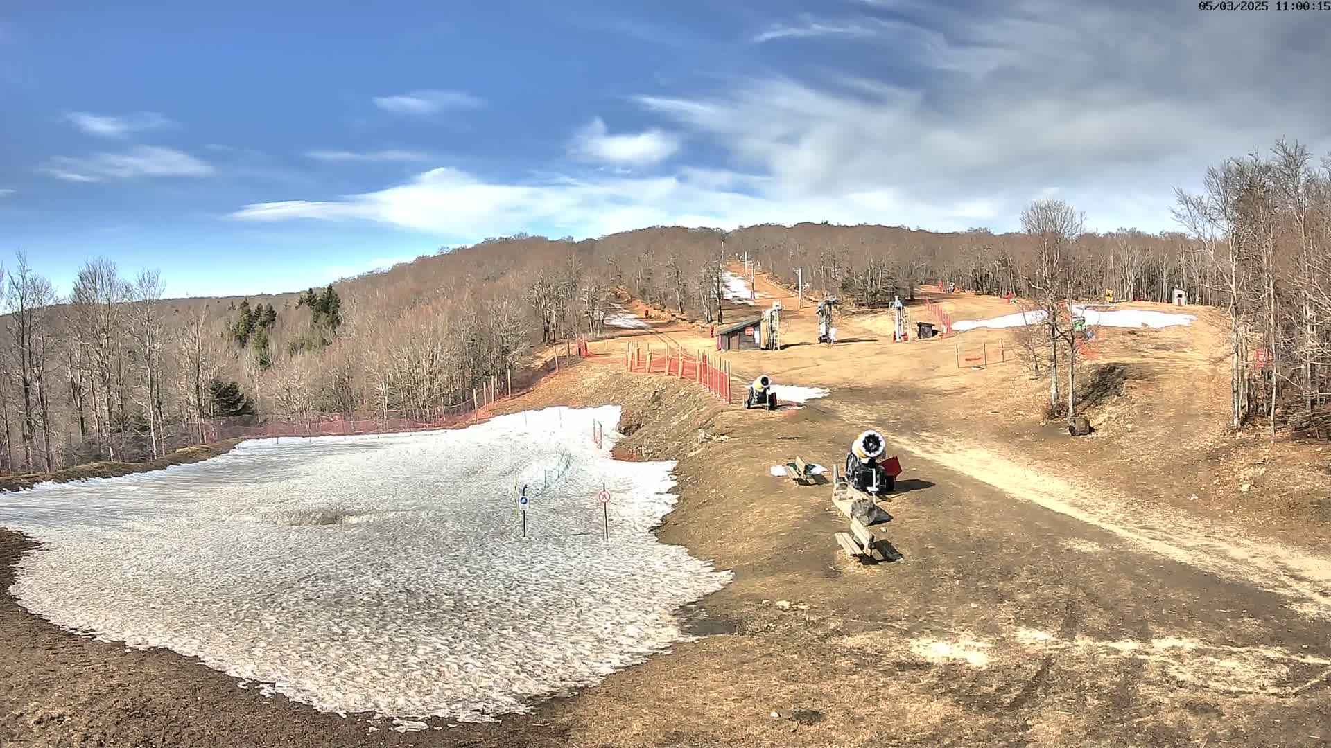 A mostly bare ski slope with patches of snow, snowmaking machines, and a mostly leafless forest in the background under a mostly sunny sky.