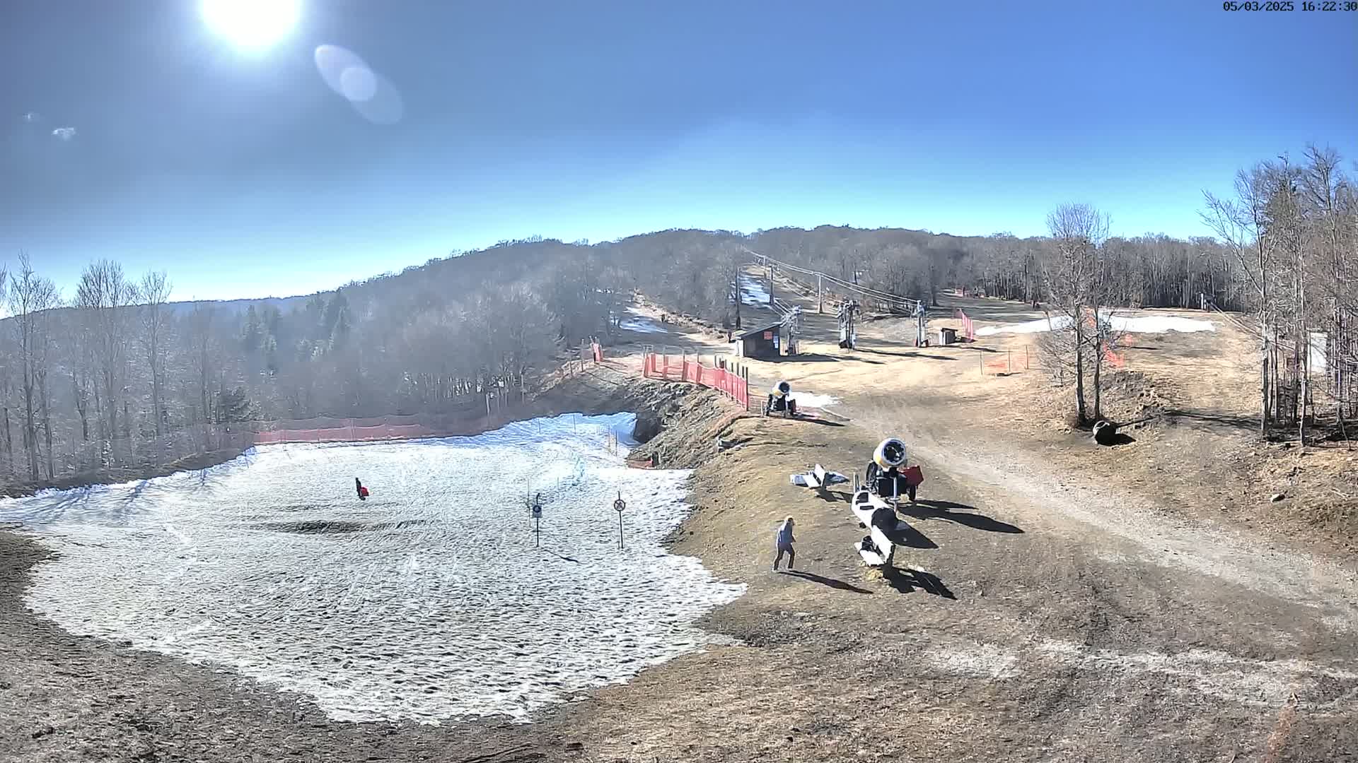 A sunny day at a ski resort shows a partially snow-covered slope with snowmaking machines and a few people present, surrounded by bare trees and brown ground.