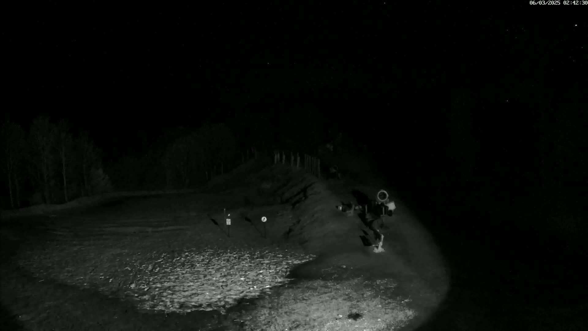 A nighttime, low-light image shows a dark, sparsely vegetated area with a dirt embankment and some equipment near a small, possibly fenced area.