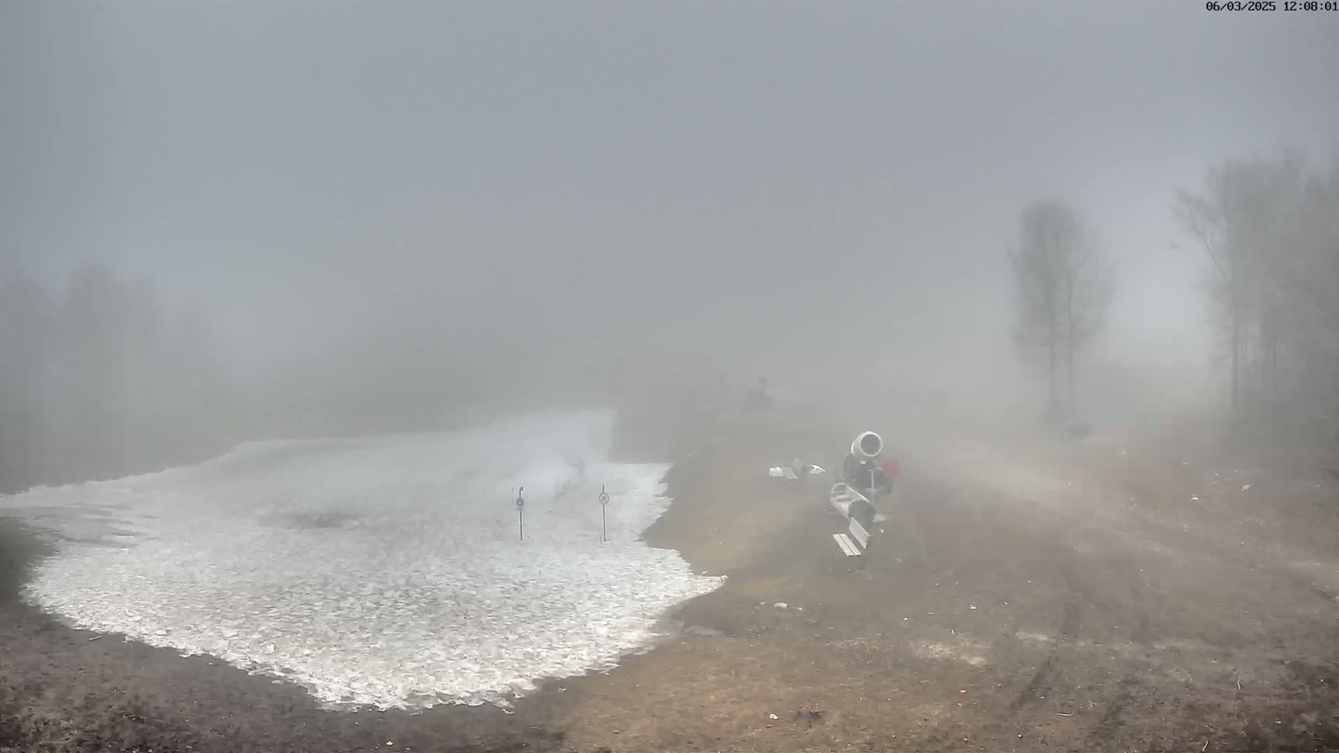 A foggy landscape shows a patch of snow next to a dirt road, with snowmaking equipment and a few people visible in the distance.
