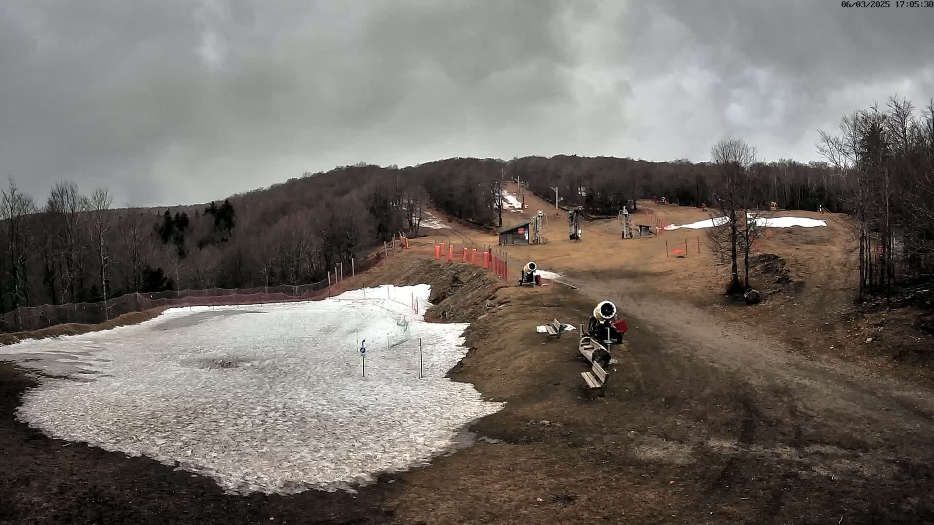 Under a cloudy sky, snowmaking machines sit on a mostly bare ski slope with patches of snow, surrounded by leafless trees.
