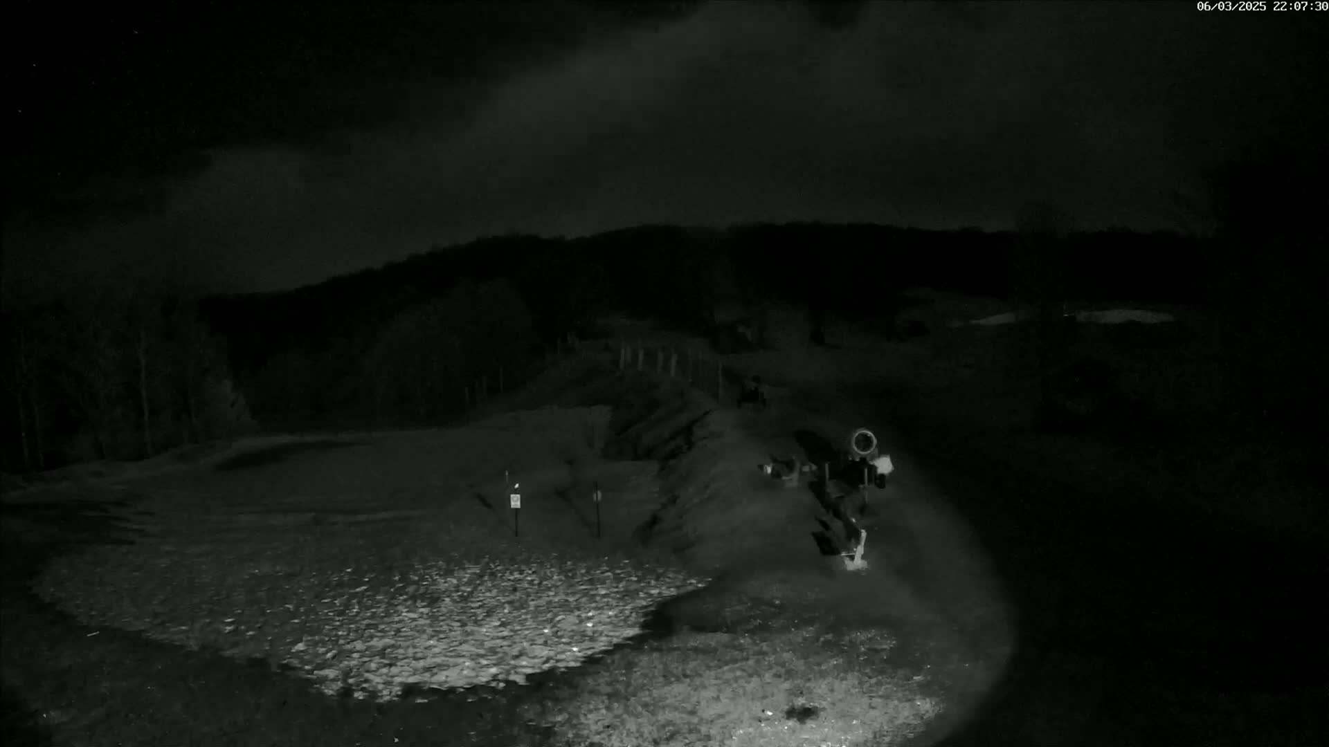 A nighttime, low-light image shows a gravel area with a person near a piece of equipment in front of a dark treeline under a cloudy sky.