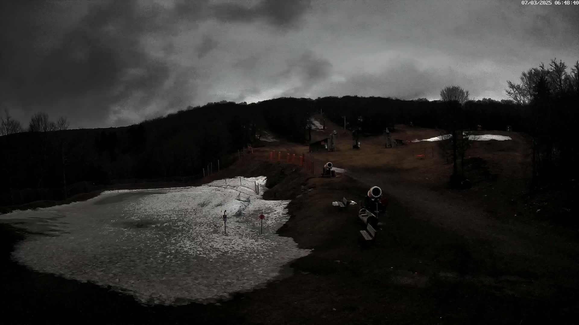 A snowmaking operation is underway on a ski slope under a dark, cloudy sky.