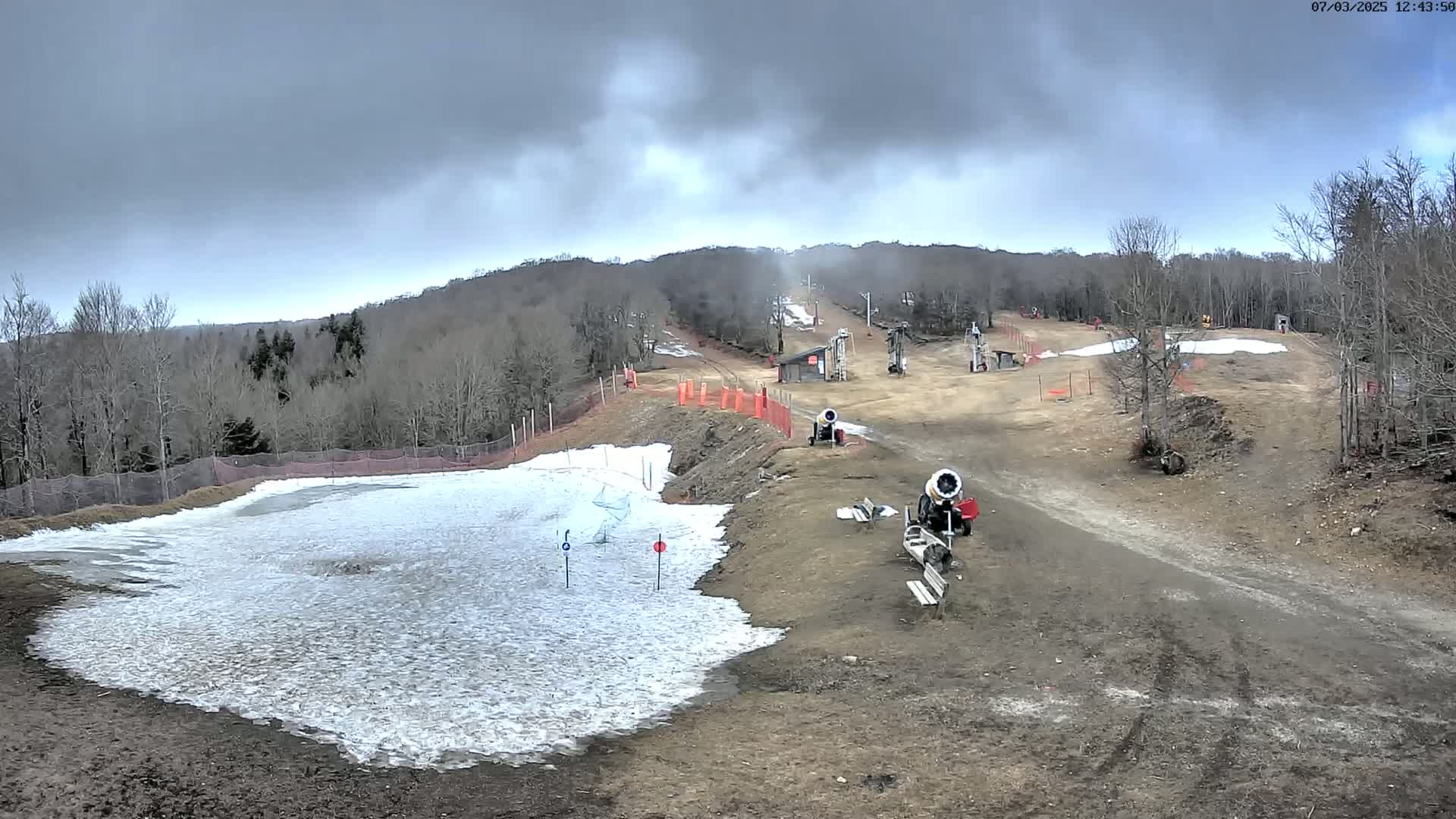 A snowmaking operation is underway at a ski resort under an overcast sky, with snow guns visible near a partially snow-covered slope.