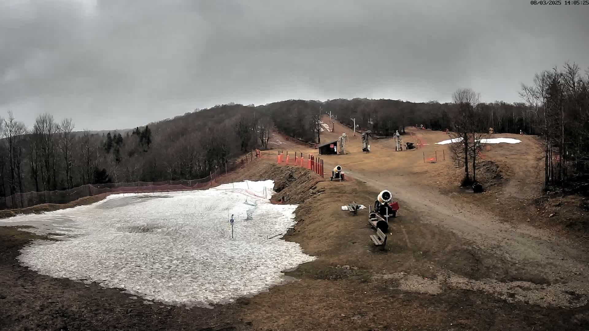 Under a cloudy sky, a snowmaking operation is visible on a sparsely snow-covered ski slope with snow guns and a small amount of snow on the lower slopes.