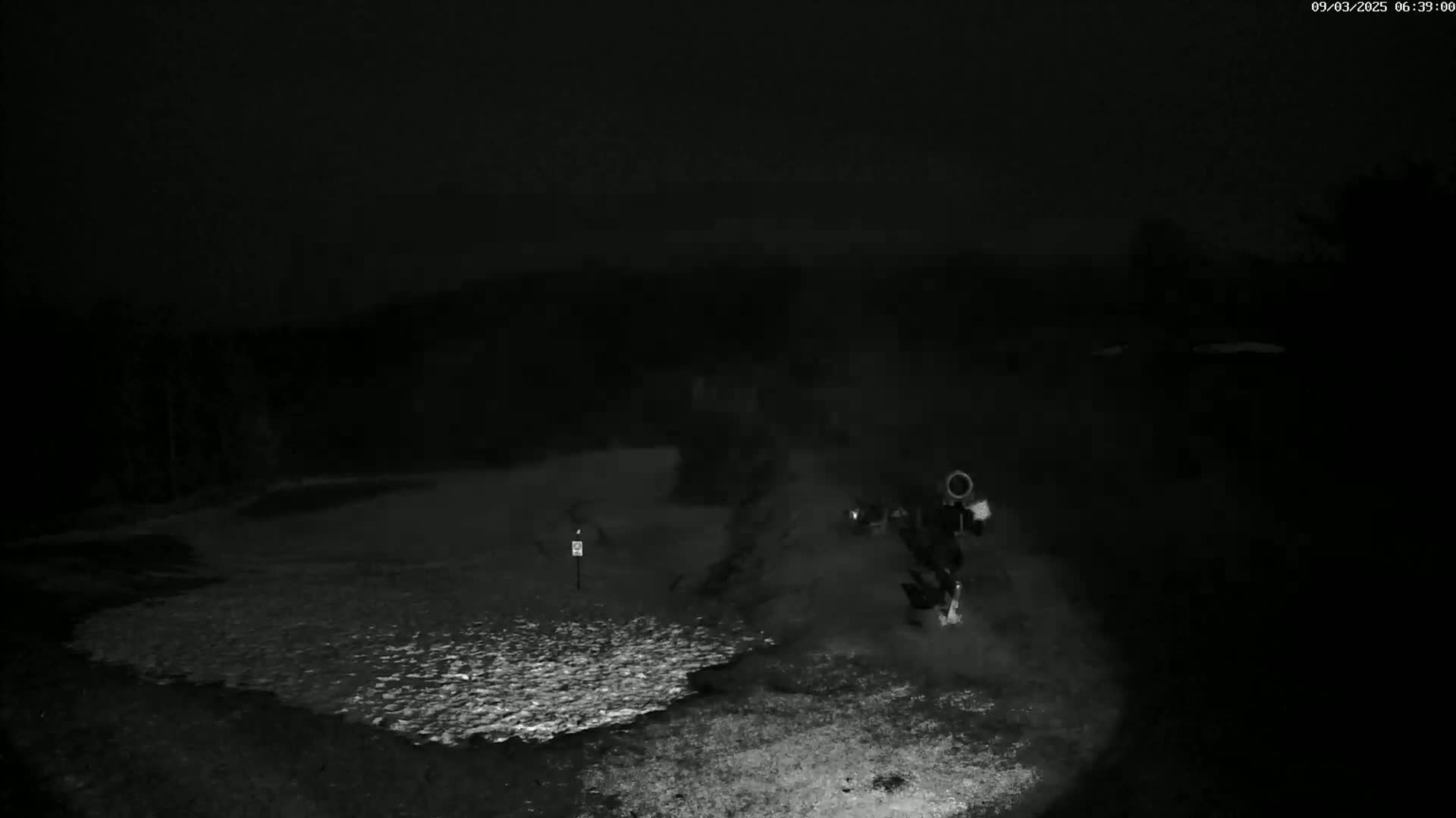 A nighttime, low-light image shows a snow-covered ground with a person and object in the middle-right, and a small marker in the left-middle ground.