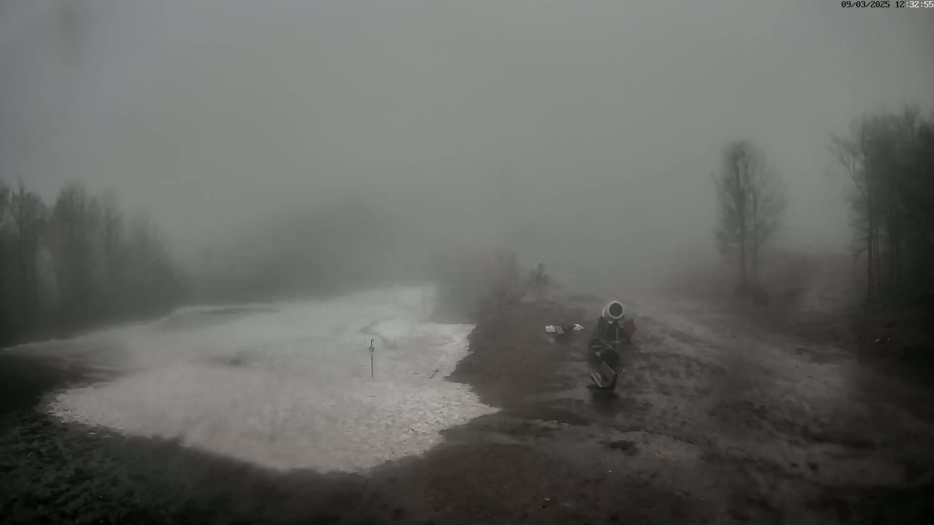 A snowmaking machine sits on a muddy hilltop overlooking a partially snow-covered area, all enveloped in thick fog.