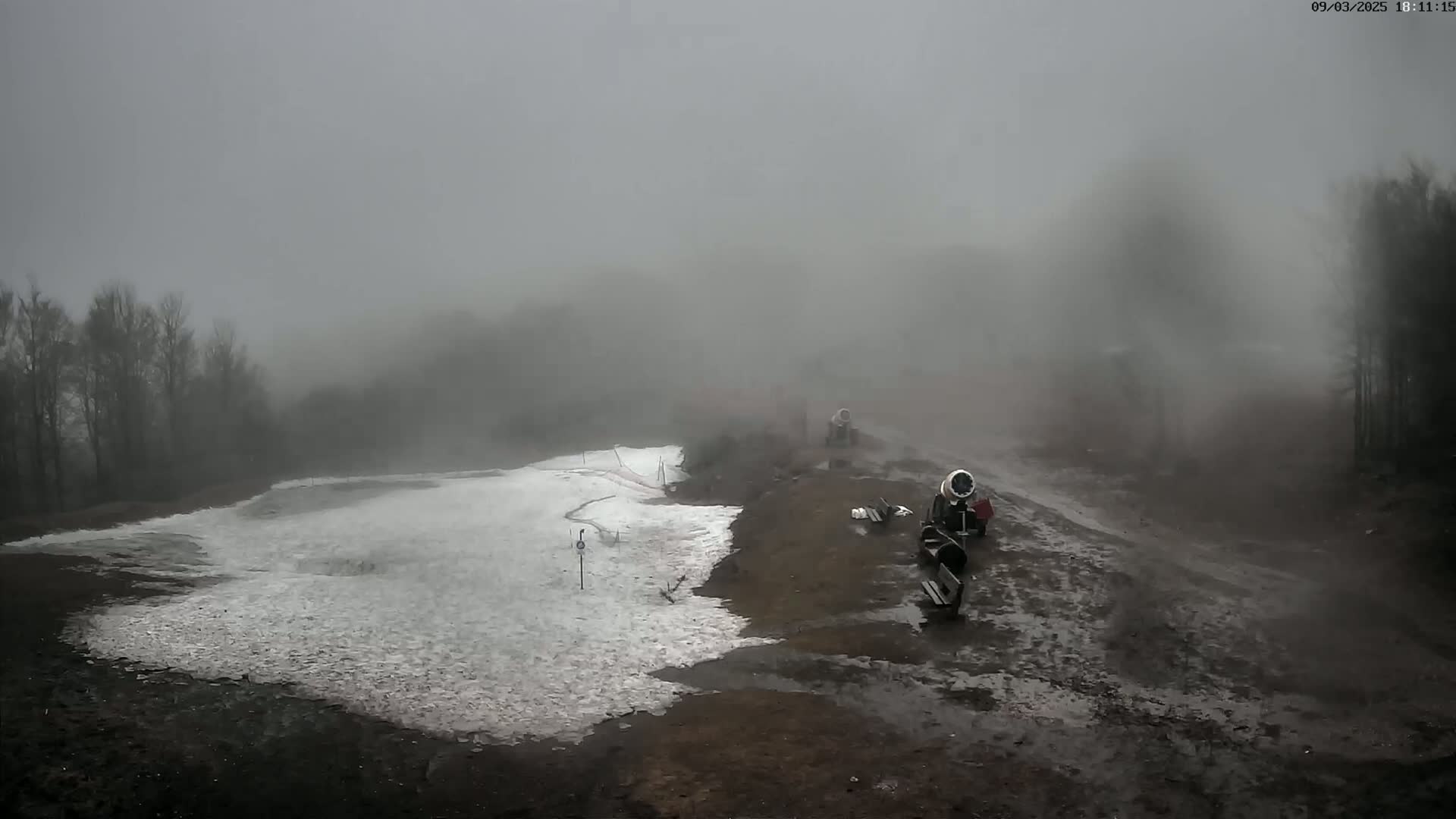 A foggy, overcast day reveals a partially snow-covered ski slope with snowmaking machines and muddy ground.