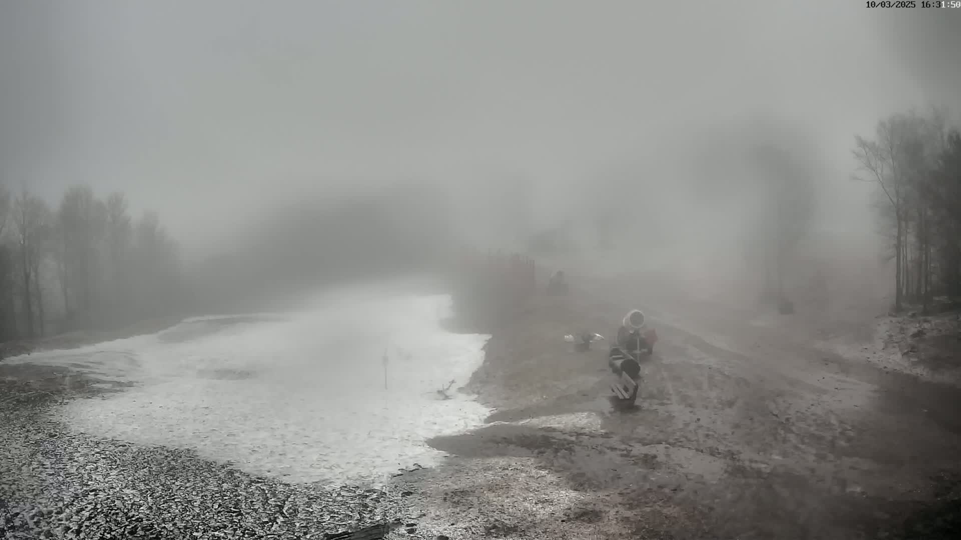 A foggy, snowy landscape shows a muddy area next to a snow-covered slope, with a few people and objects visible in the hazy distance.