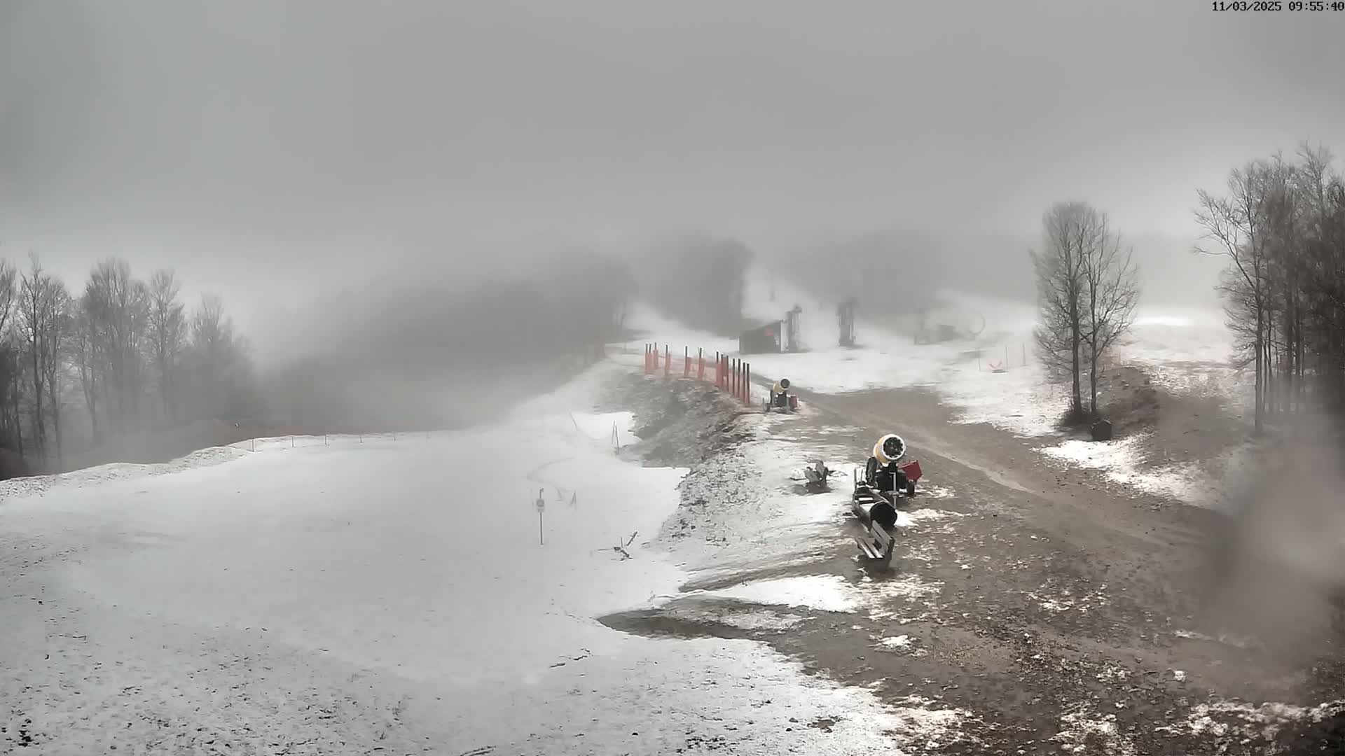 A snow-making machine sits on a partially snow-covered ski slope in foggy conditions, with bare trees visible in the background.