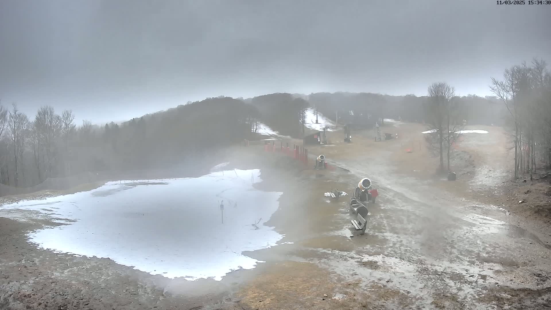 A misty, overcast day at a ski resort shows a partially snow-covered slope with snowmaking machines and bare trees in the background.