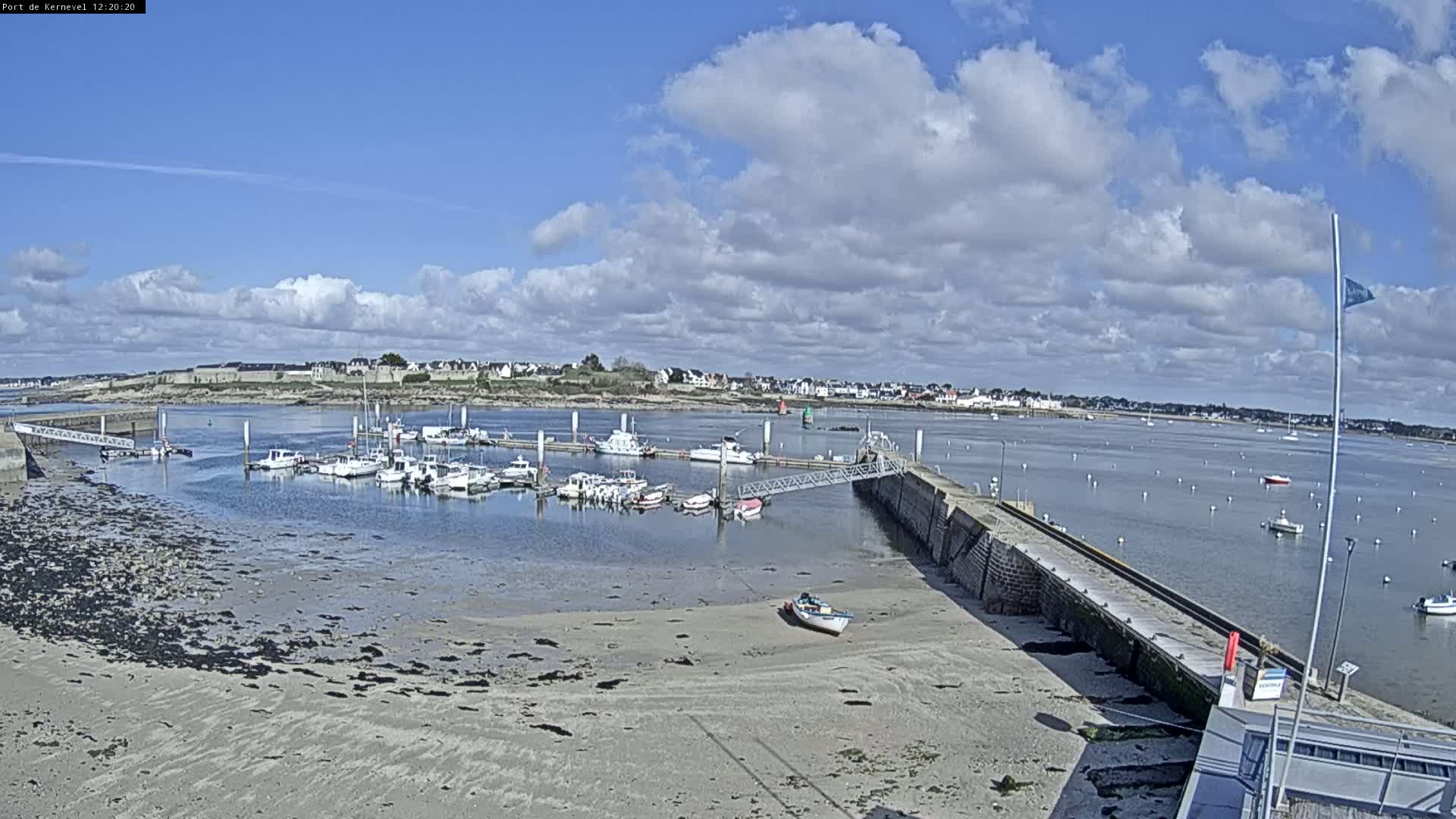 A mostly sunny day reveals a harbor filled with boats, a sandy shoreline, and a small town in the distance.