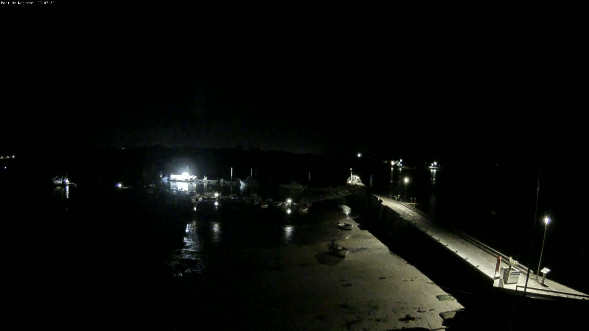 A nighttime, high-angle view of a harbor with boats docked, a long pier extending into the water, and a few lights illuminating the scene.