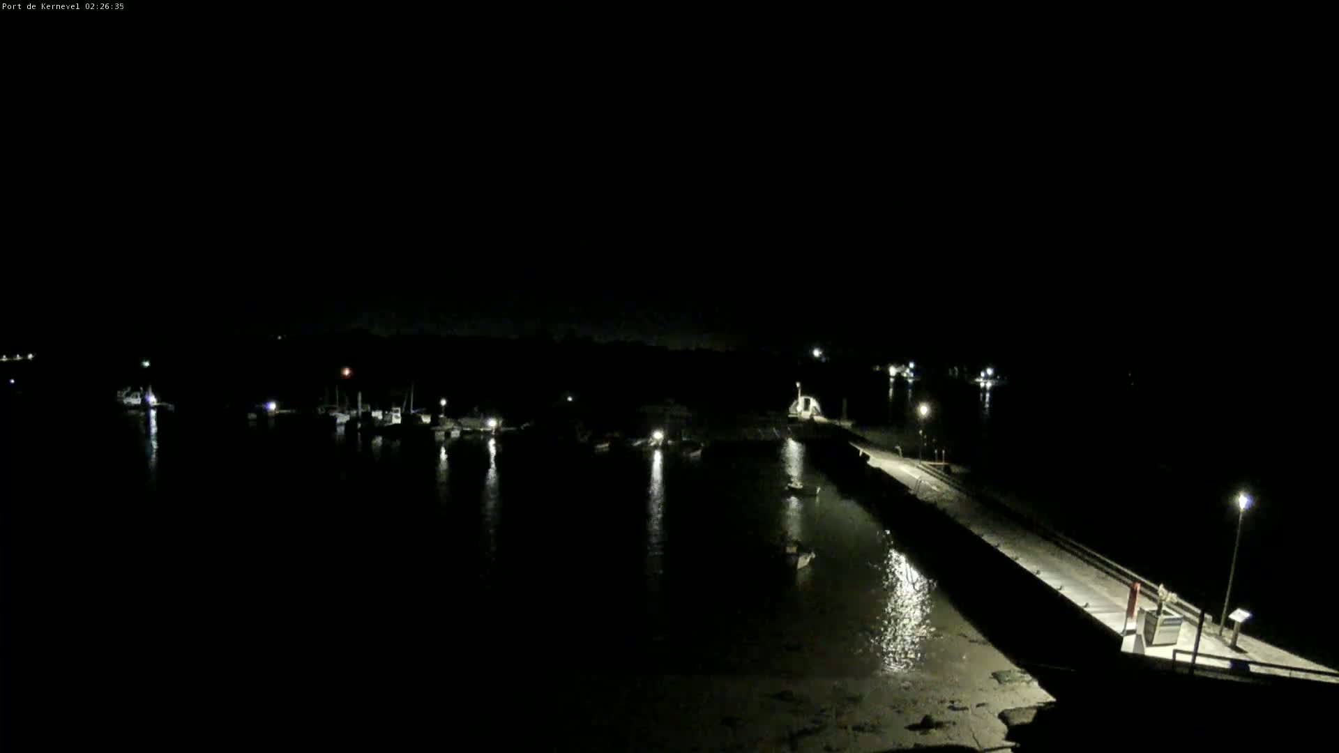 A dark nighttime harbor scene shows several boats moored along a pier, with calm water reflecting lights.