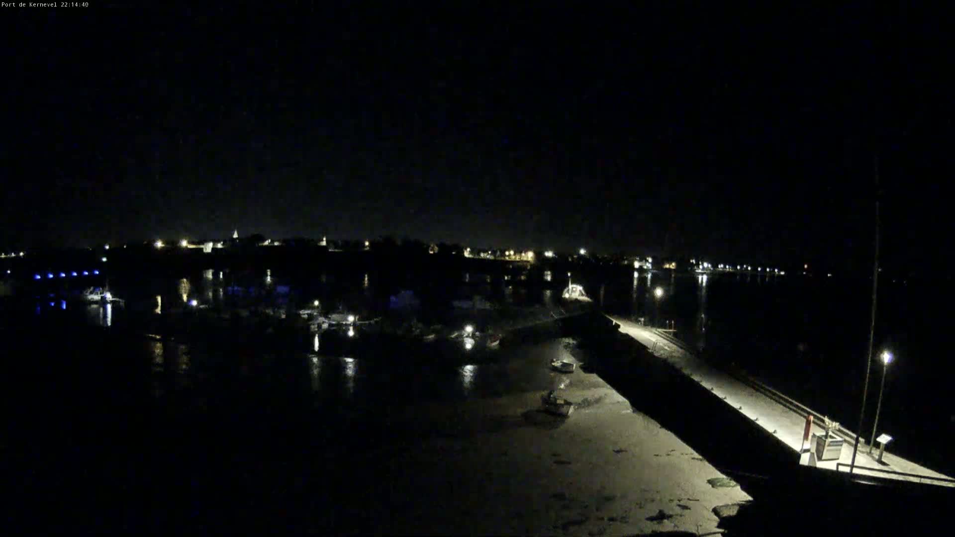 A nighttime view of a harbor with several boats and a long pier extending into the water under a clear, dark sky.