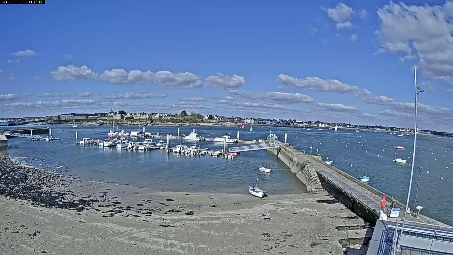 A marina filled with boats is situated on a calm, sunny day with partly cloudy skies, next to a sandy shore and a town in the distance.