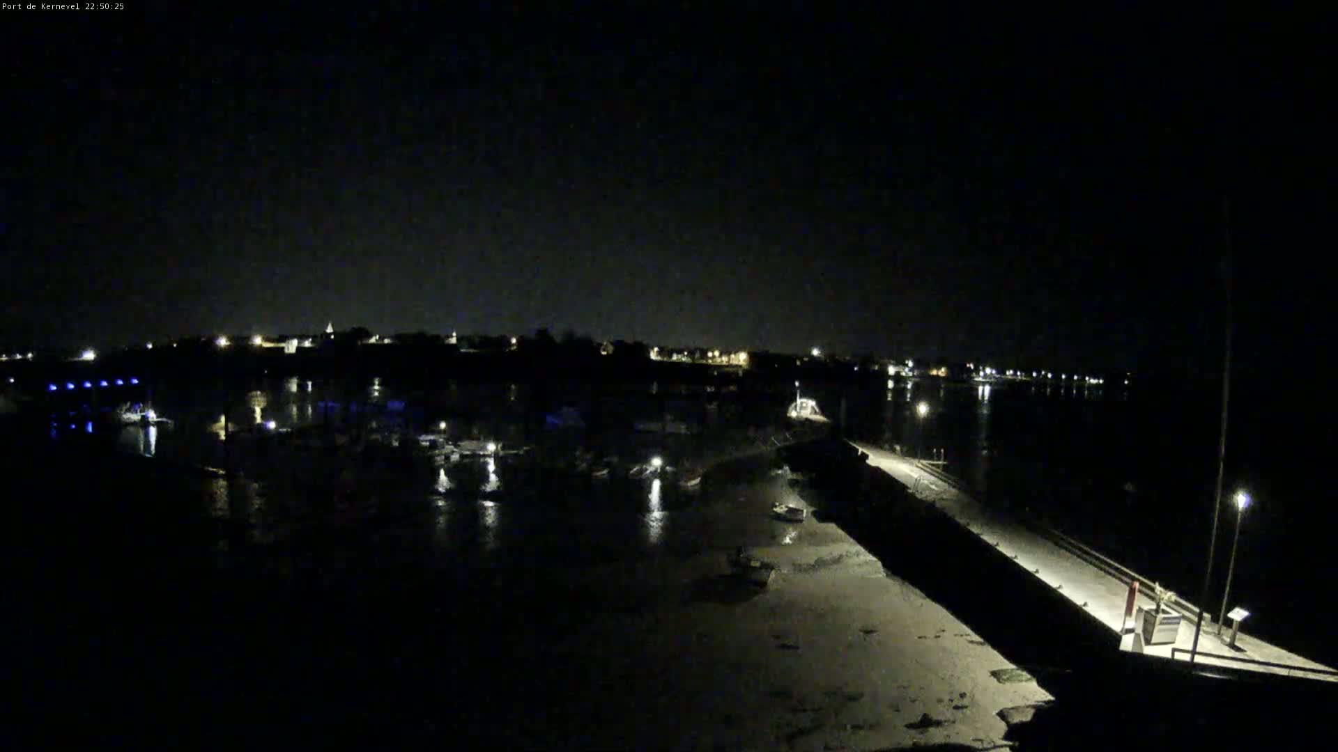 A nighttime, low-light view of a harbor with several boats docked, a long, dark wall extending into the water, and a small town faintly visible in the distance under a clear, dark sky.