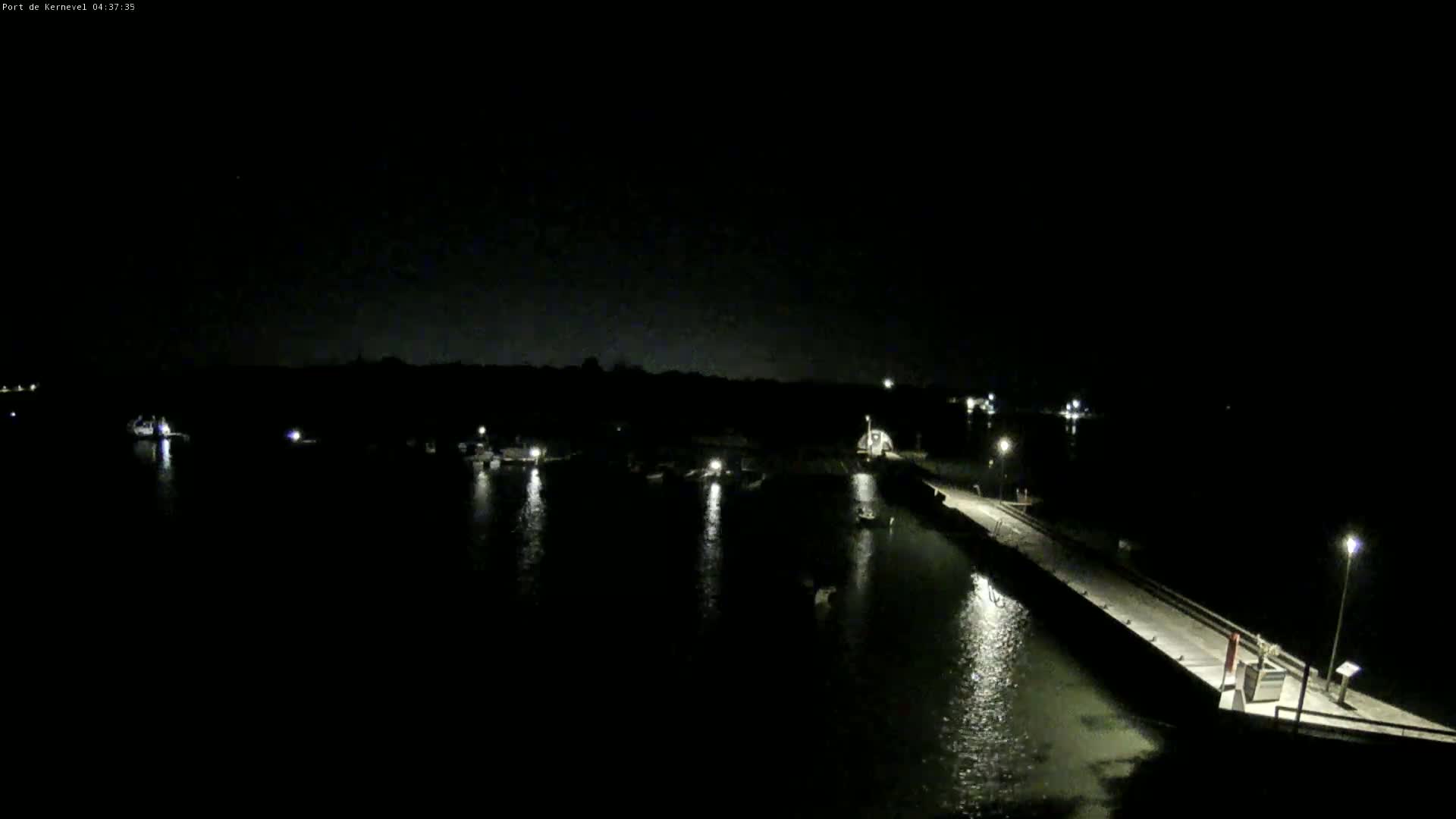 A nighttime view of a harbor with several boats, a long dock, and dimly lit land.