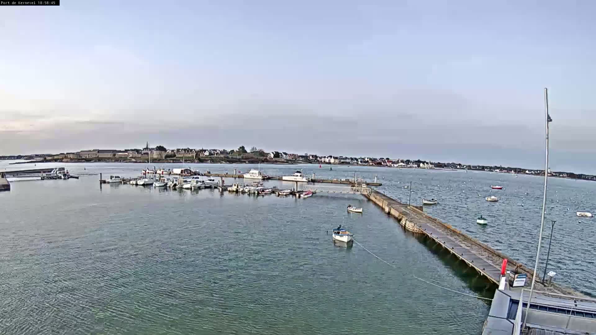 A calm body of water shows a marina filled with several boats, with a small town visible in the background under a mostly clear sky.