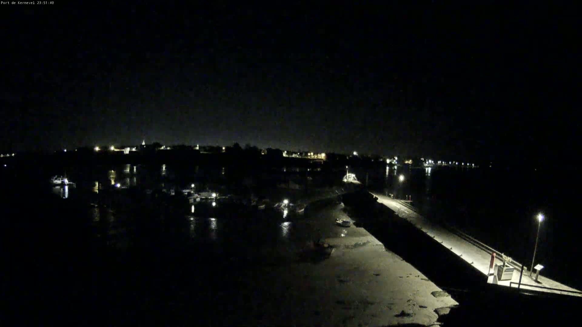 A nighttime, low-light view of a harbor with boats and a long pier extending into the water under a clear, dark sky.
