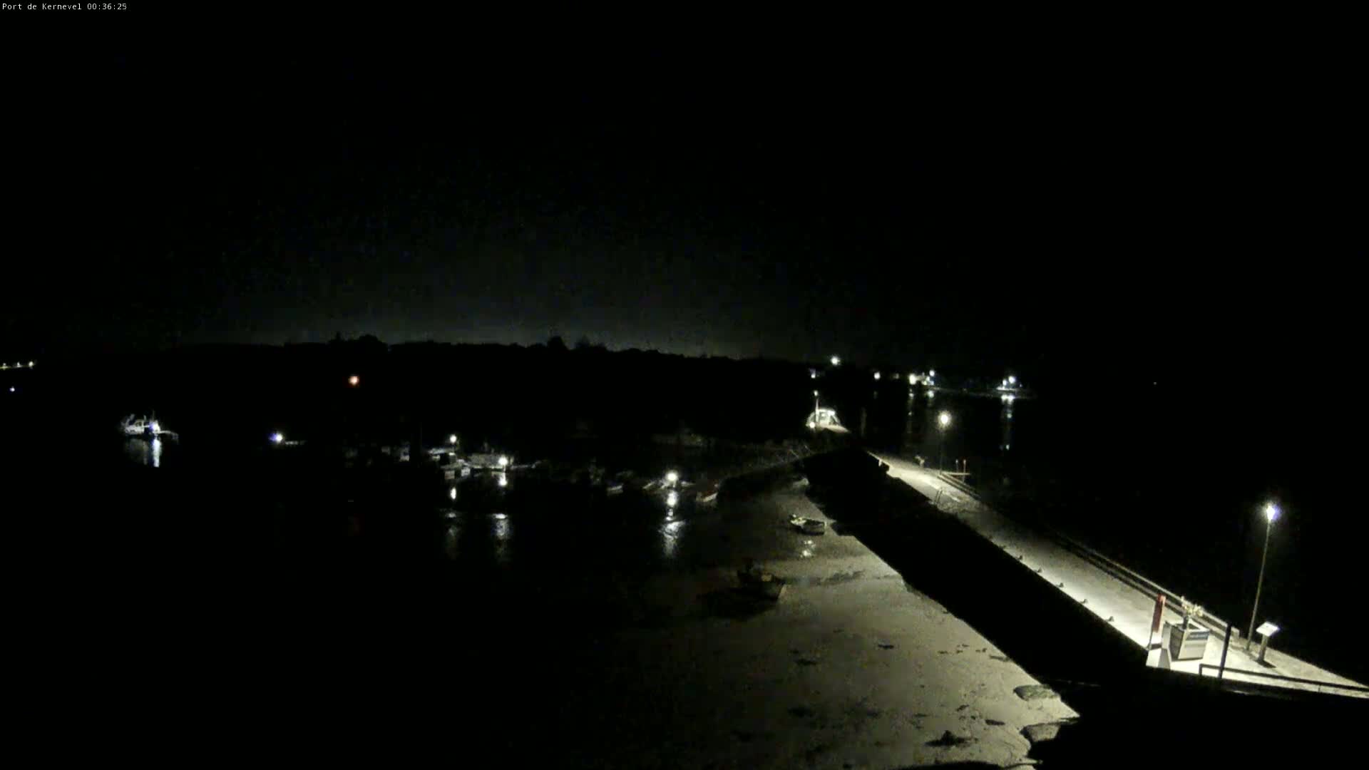 A nighttime, high-angle view shows a dark harbor with sparsely lit boats and a long, dark pier extending into the water.