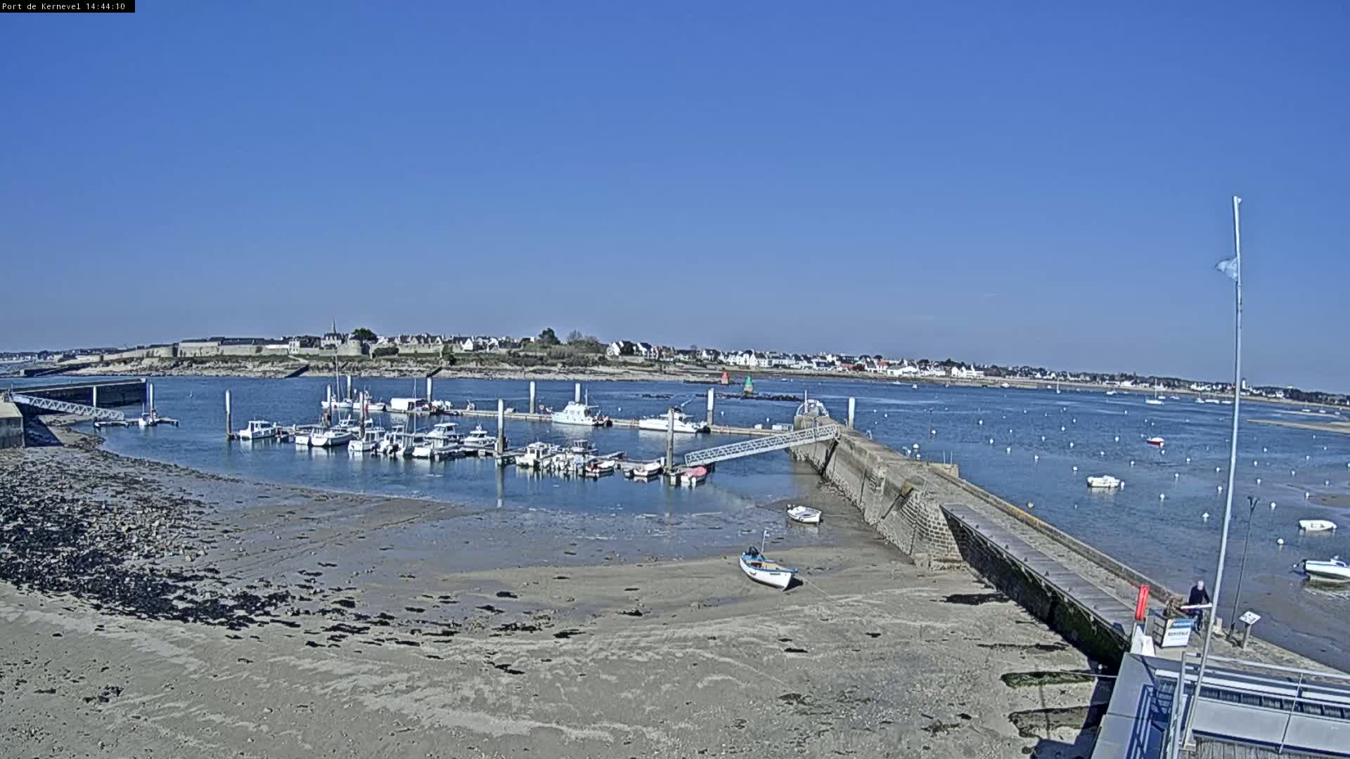 A small harbor with numerous boats is situated on a sandy shore under a clear blue sky.