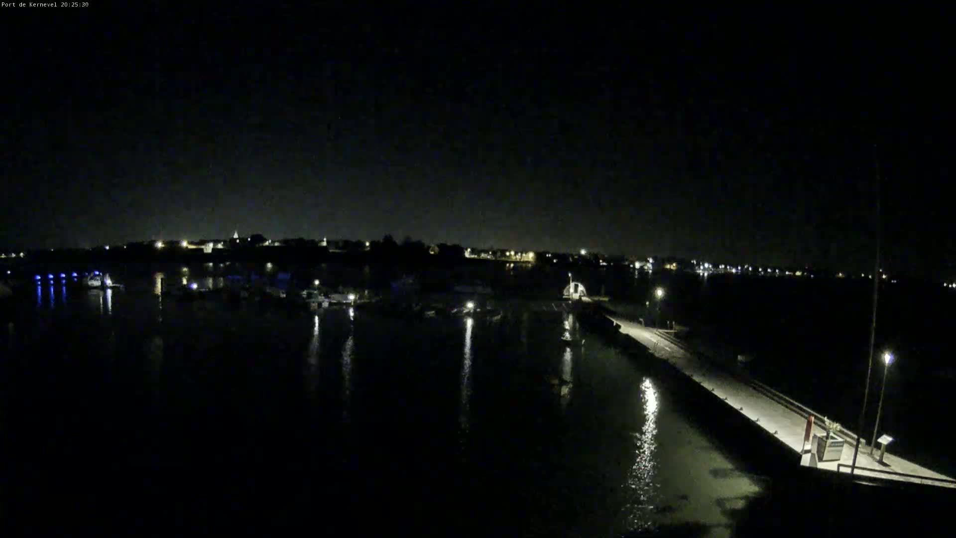 A nighttime, low-angle view of a calm harbor with docked boats and a pier extending into the water, under a dark sky.