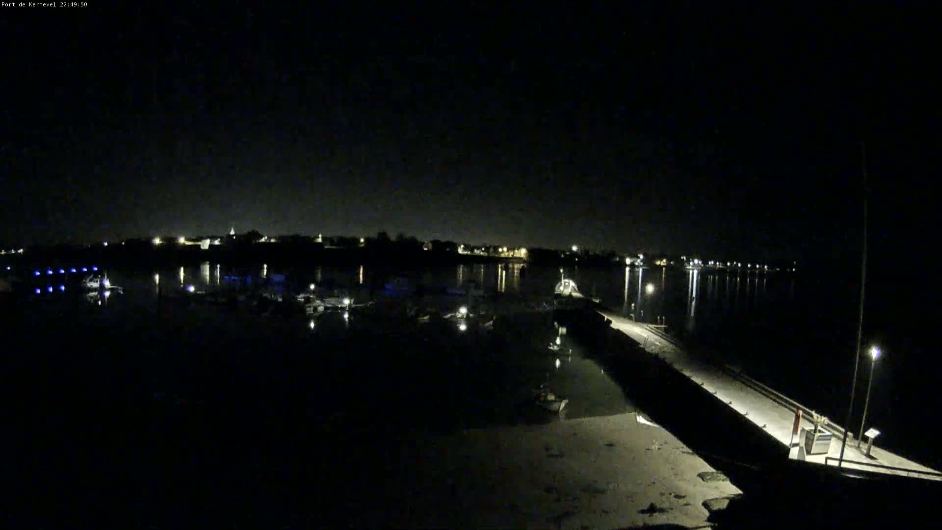 A nighttime view of a harbor with boats docked, a pier extending into the water, and a town visible in the distance under a clear, dark sky.