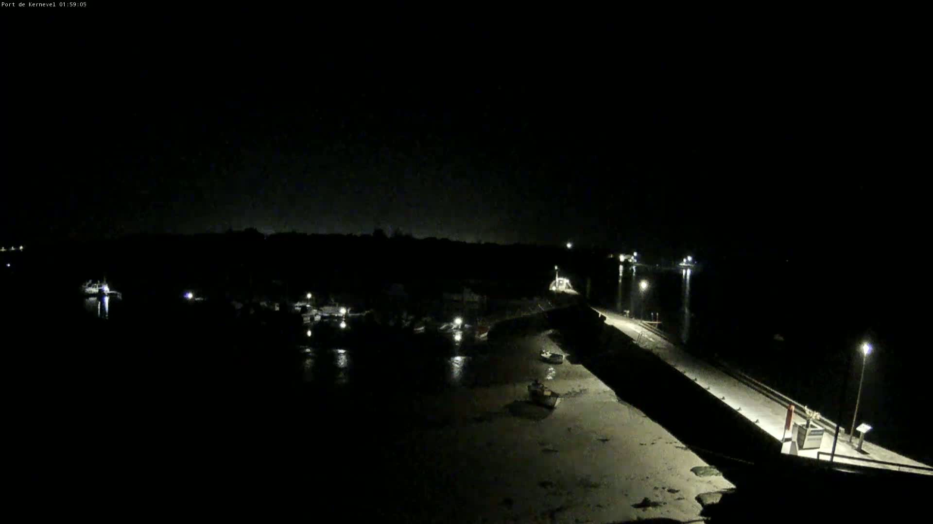 A nighttime view of a harbor with several boats docked, a long pier extending into the water, and some land visible in the background under a dark sky.