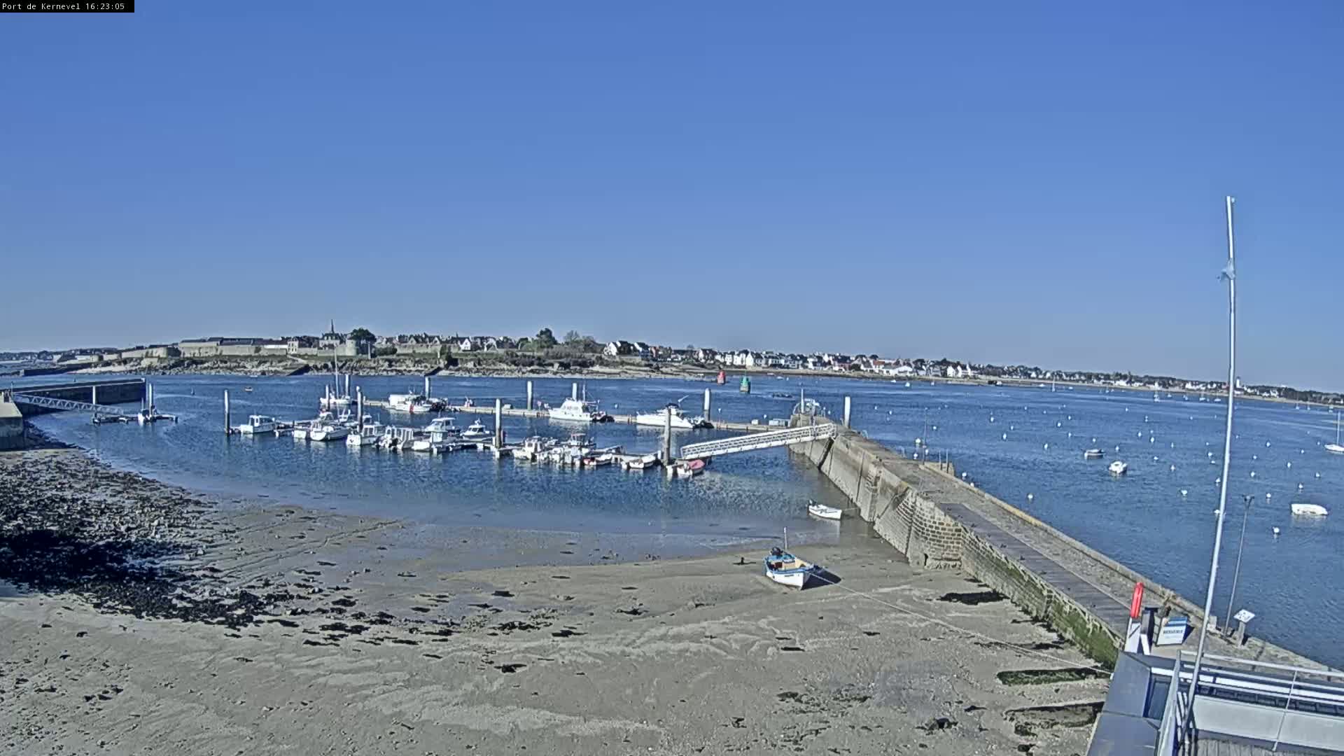 A marina filled with boats sits on a calm, sunny day next to a sandy shore and a small town in the distance.