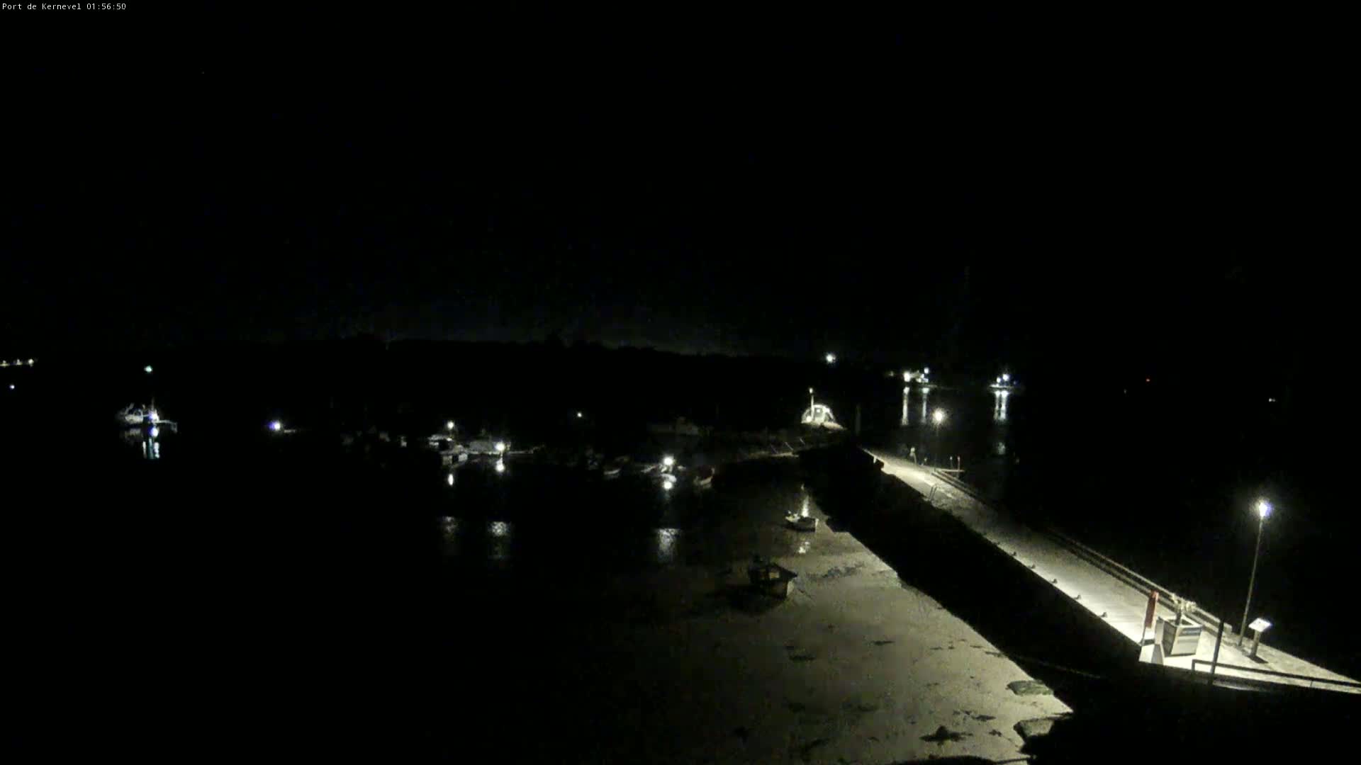 A nighttime view of a harbor with several boats moored, a long pier extending into the water, and the surrounding land mostly dark under a clear sky.