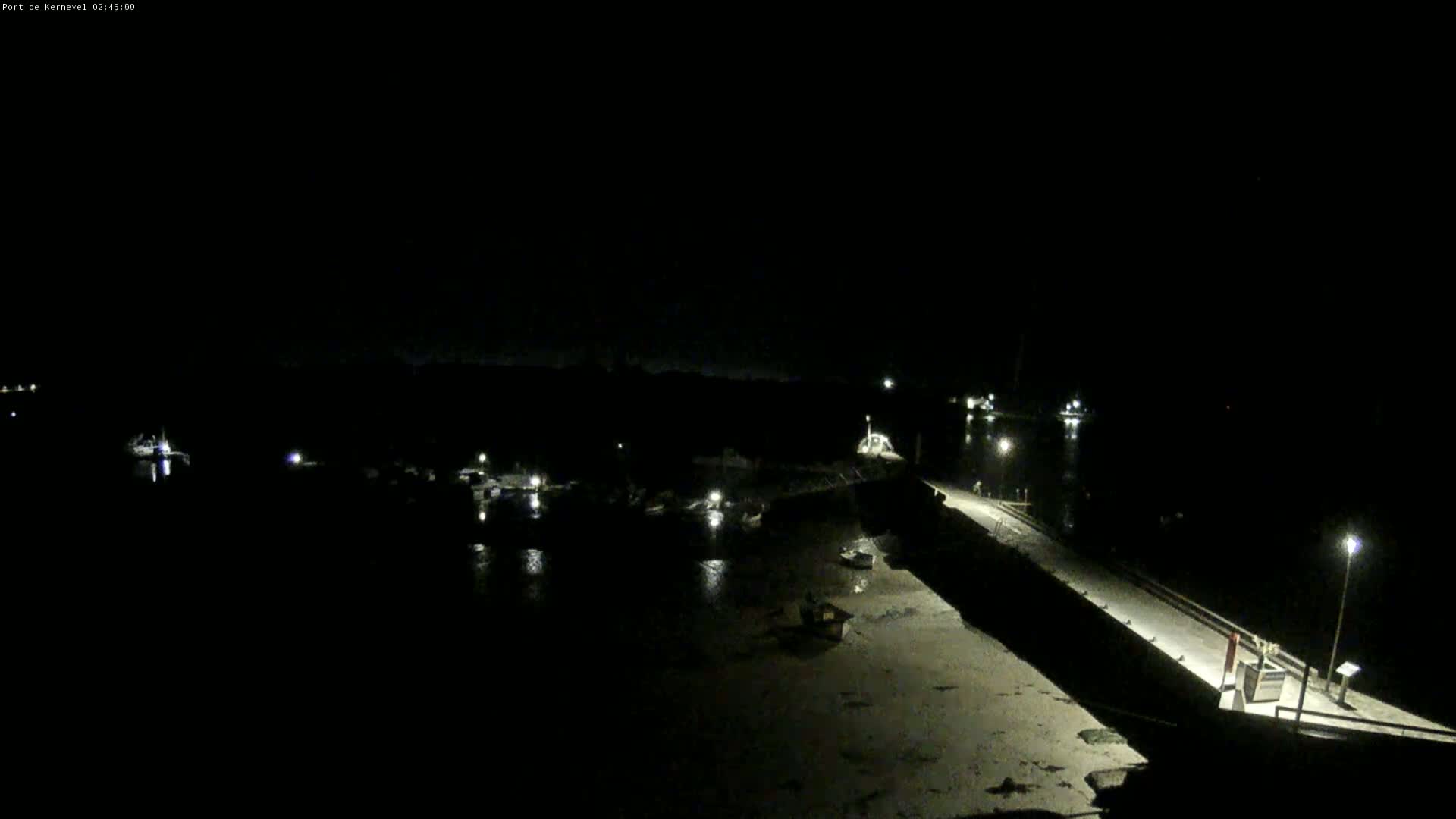 A nighttime, low-light view of a harbor with several boats moored near a long pier extending into the water.