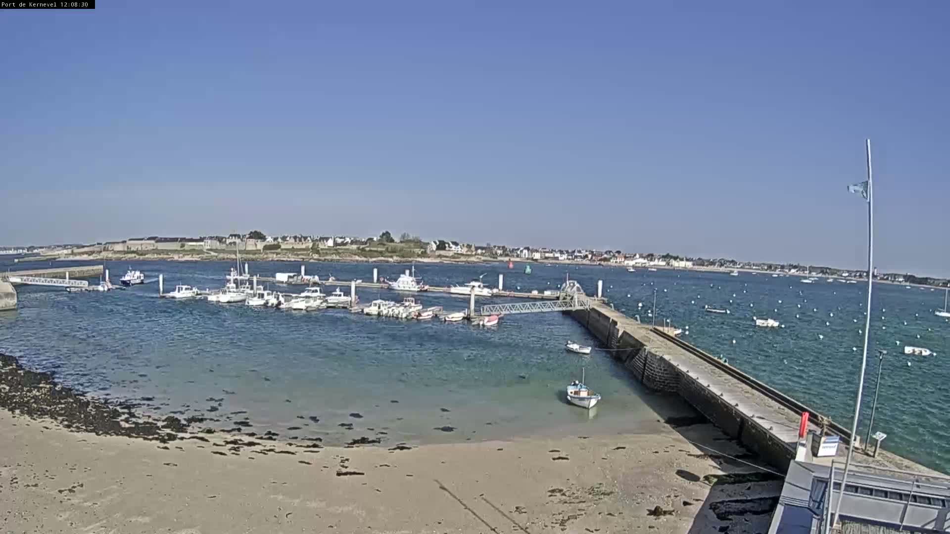 A small harbor filled with numerous boats is nestled on a sandy shore, under a clear, sunny sky.