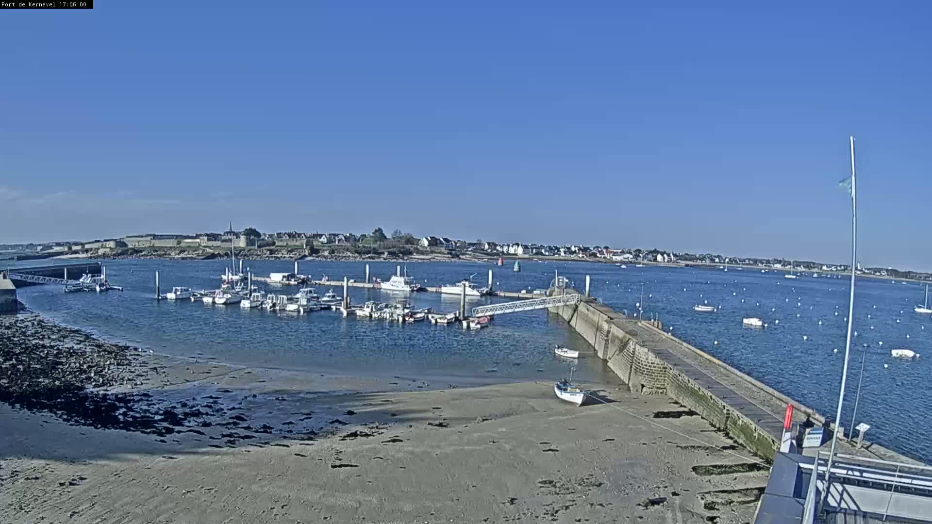 A marina filled with numerous boats is situated on a calm, sunny day, with a sandy shoreline and a small town visible in the distance.