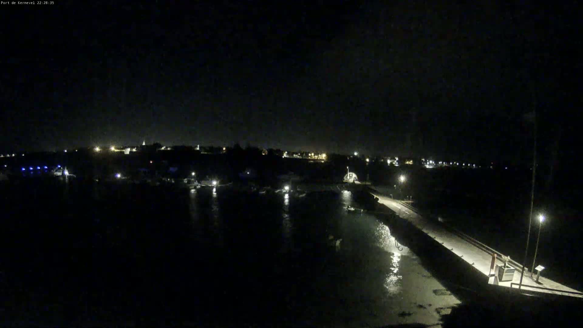 A nighttime view of a dark harbor with a pier extending into the water, illuminated by scattered lights along the shoreline and pier.