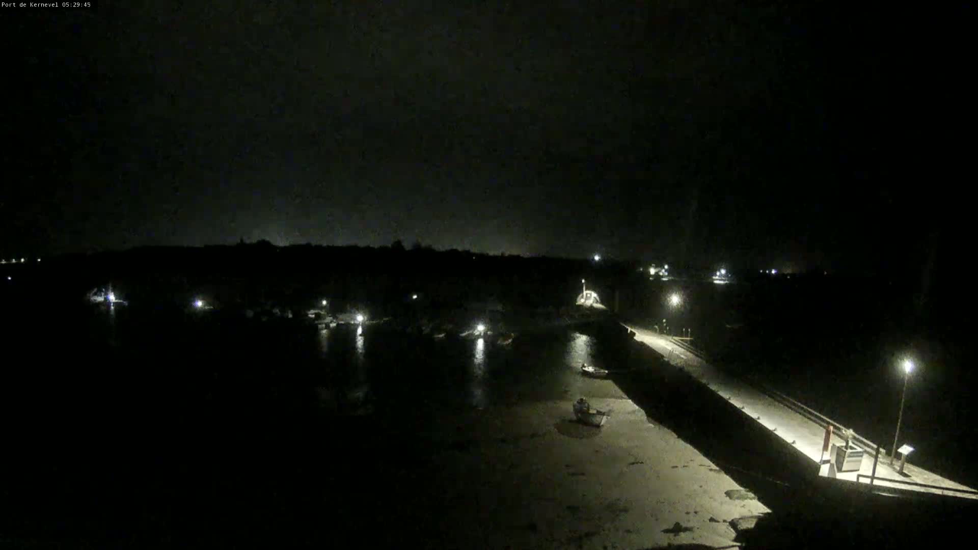 A dark nighttime scene shows a harbor with several boats and a long pier extending into the water, under a mostly cloudy sky.