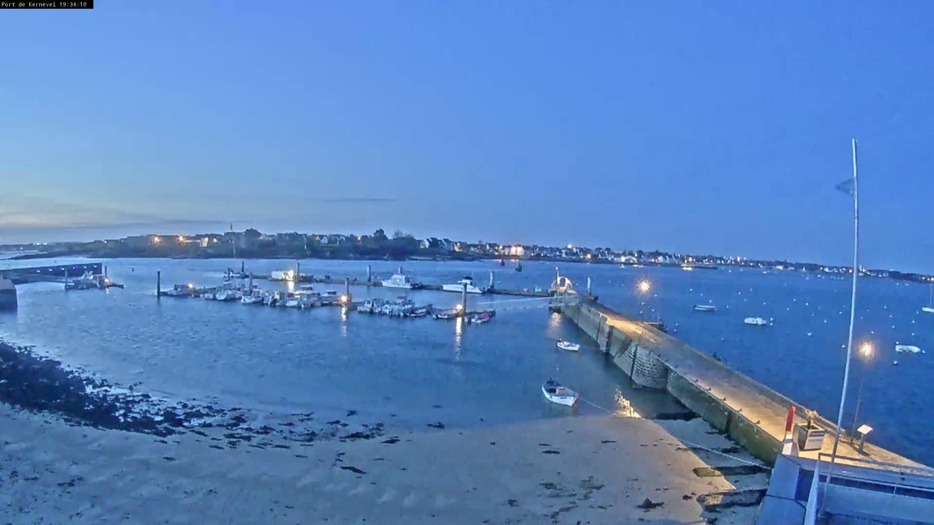 Under a twilight sky, a harbor filled with moored boats is viewed from a pier extending into calm water, with a town visible in the distance.