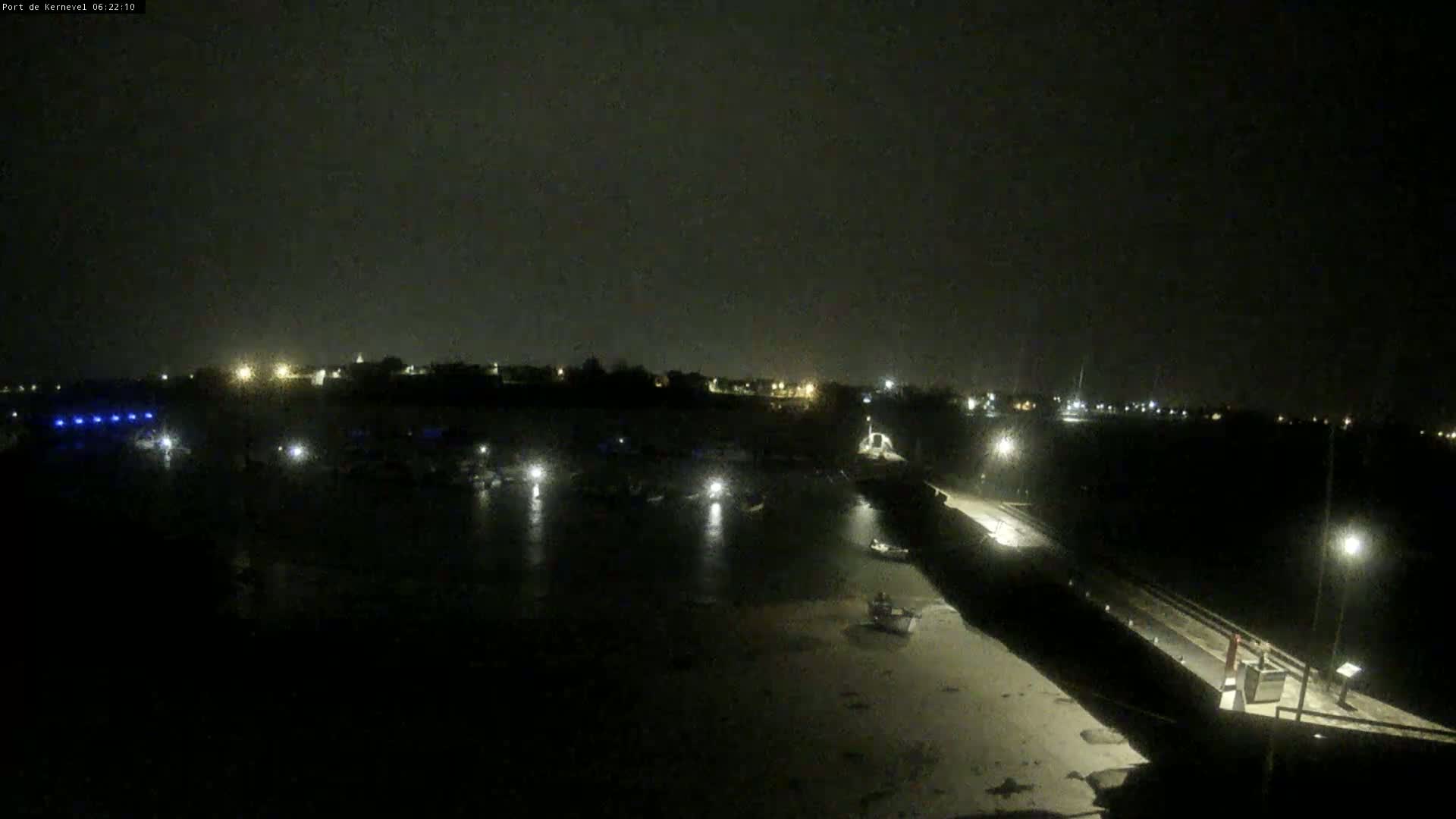 A nighttime view of a harbor with several boats, dimly lit by scattered lights under a dark, overcast sky.